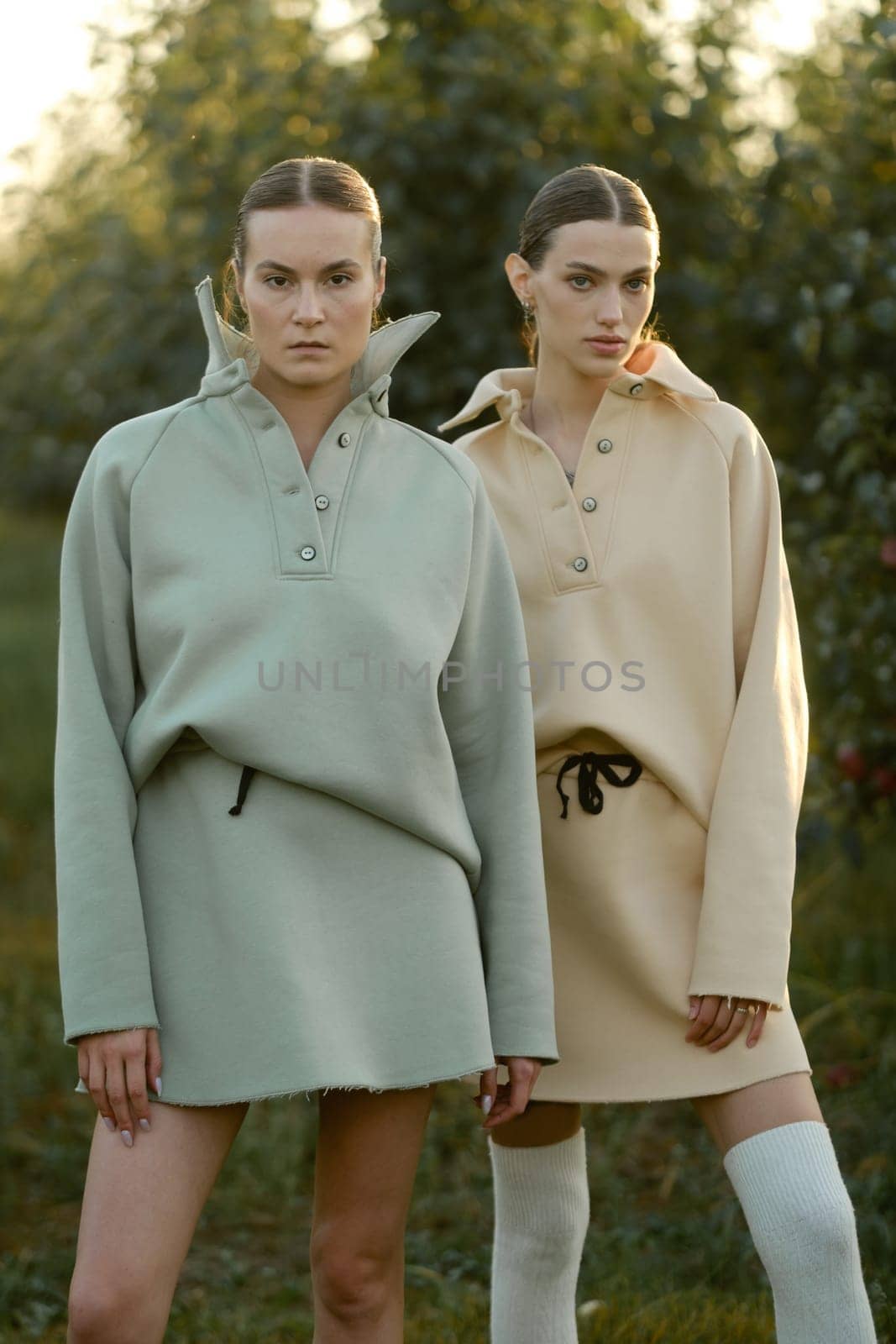 Two models in stylish tracksuits pose in apple orchard at sunset . The model is wearing a light-colored tracksuit and skirt.