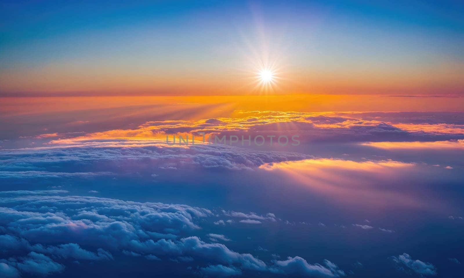 An aerial view of the setting sun casting light through the clouds. The sky is a vibrant orange, yellow, and blue.