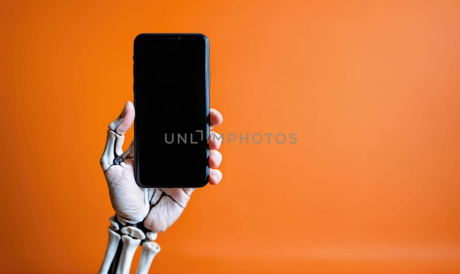 A skeletal hand holds a black smartphone vertically against an orange background. The hand is bare and the fingers are spread. The screen of the phone is black.