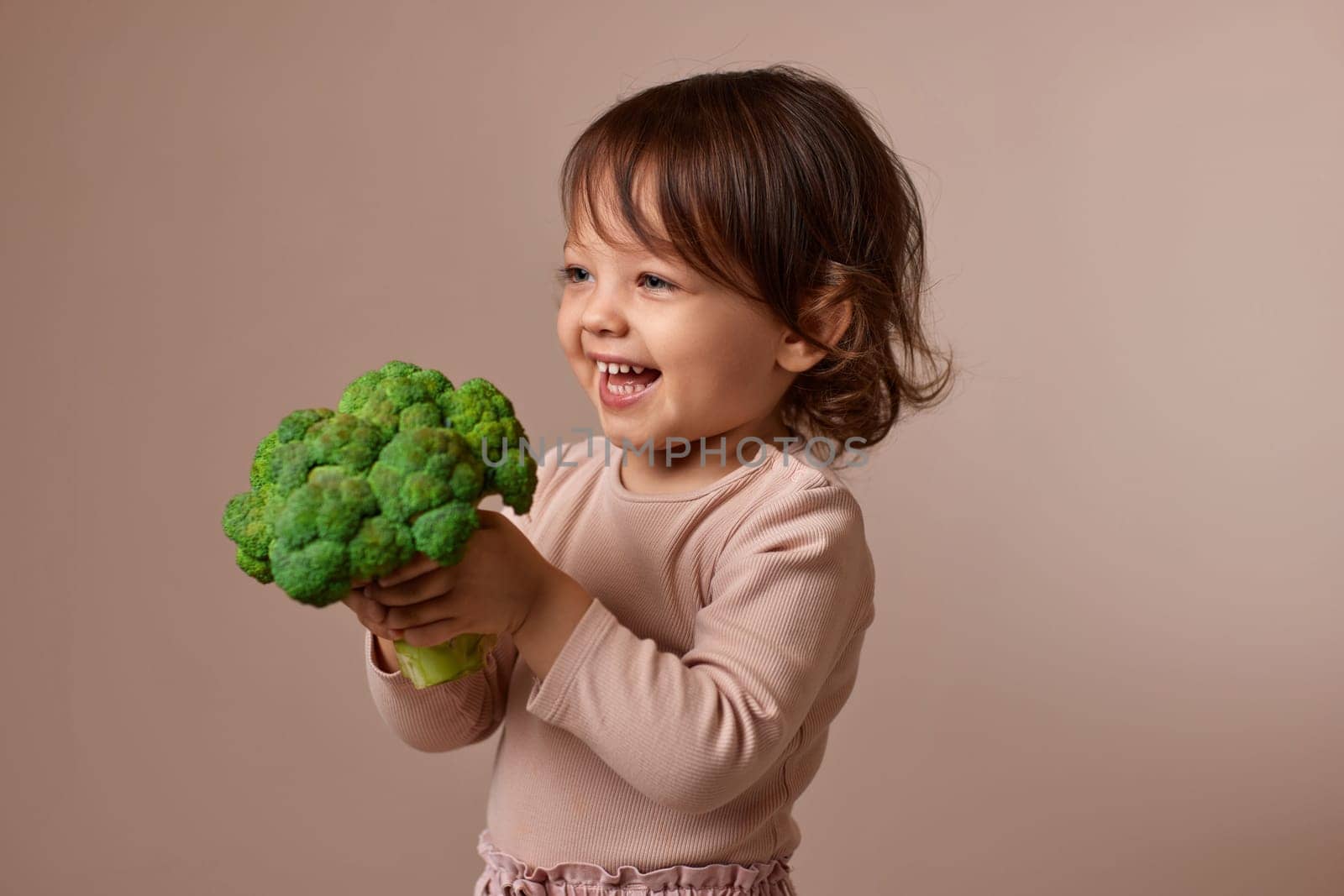 smiling child girl showing fresh green raw broccoli on beige background. healthy baby food. kid loves vegetables