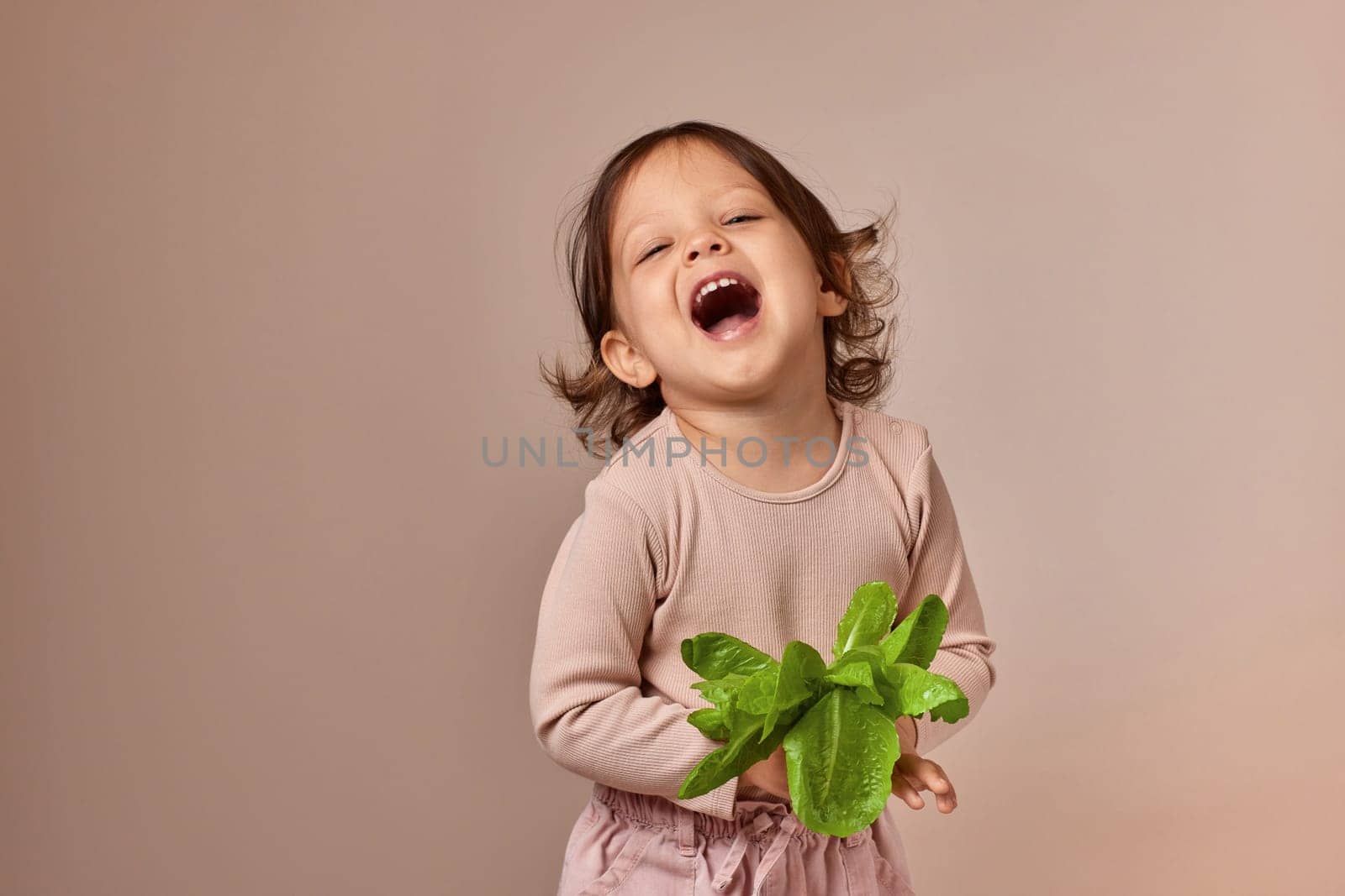 very excited smiling child girl holding fresh green salad on beige background. healthy baby food.