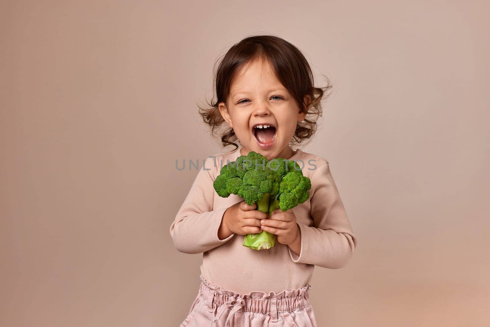 very happy child girl holding fresh green raw broccoli on beige background. healthy baby food. kid loves vegetables