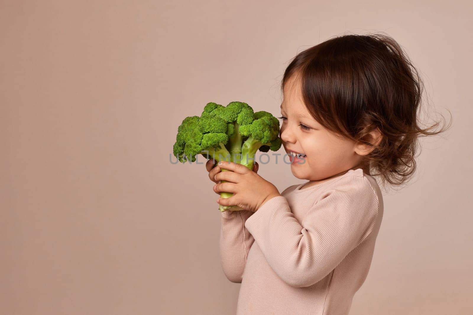 smiling little child girl holding fresh green raw broccoli on beige background. healthy baby food. kid loves vegetables