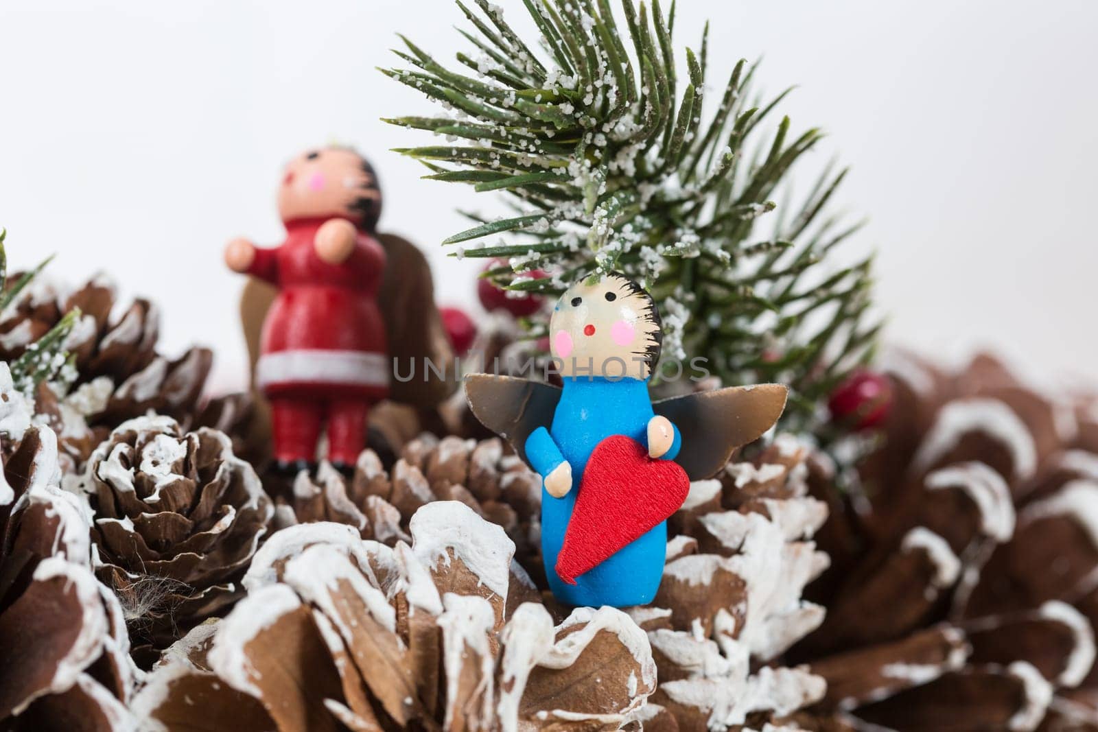 Christmas ornament featuring a small wooden angel holding a red heart, set against a festive background of pinecones and snow-dusted fir branches