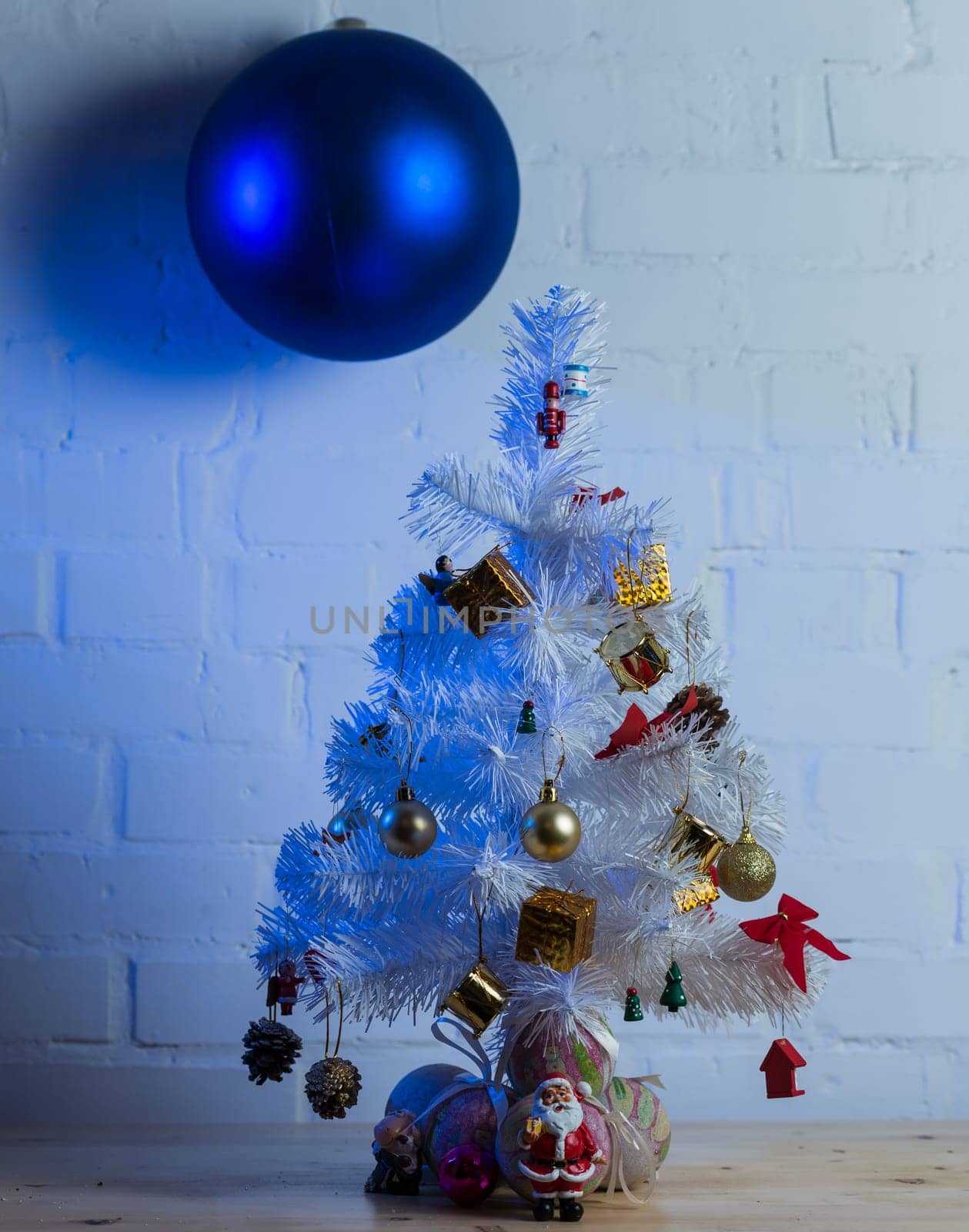 Small white Christmas tree decorated with golden and red ornaments, pine cones, and miniature Santa figurines on a wooden table against a white brick wall.