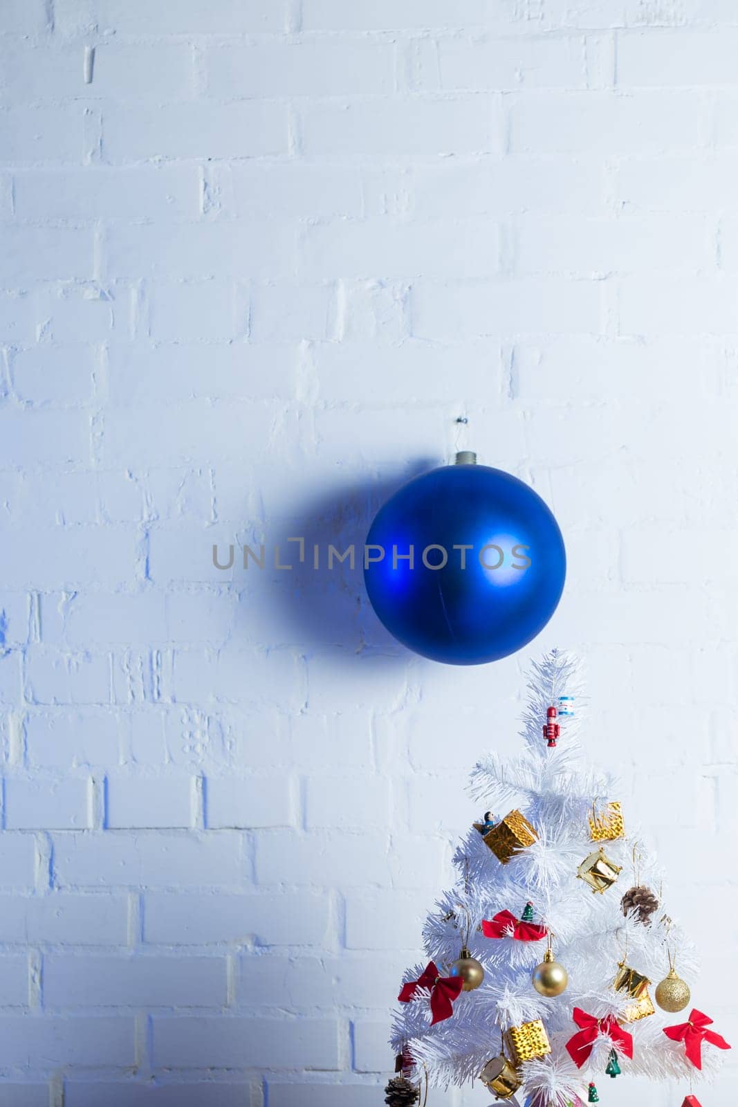 Small white Christmas tree decorated with golden and red ornaments, pine cones, and miniature Santa figurines on a wooden table against a white brick wall.