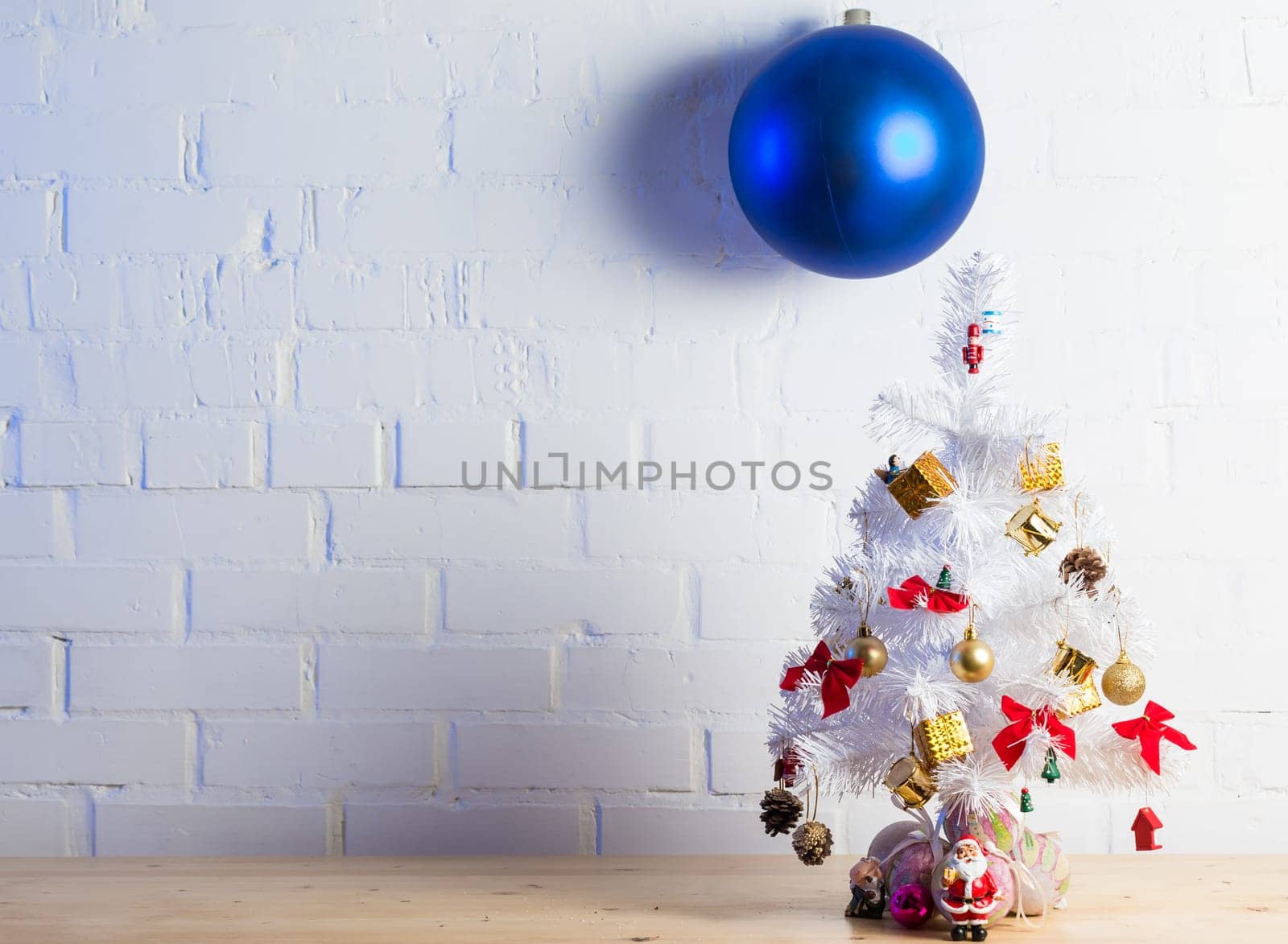 Small white Christmas tree decorated with golden and red ornaments, pine cones, and miniature Santa figurines on a wooden table against a white brick wall.