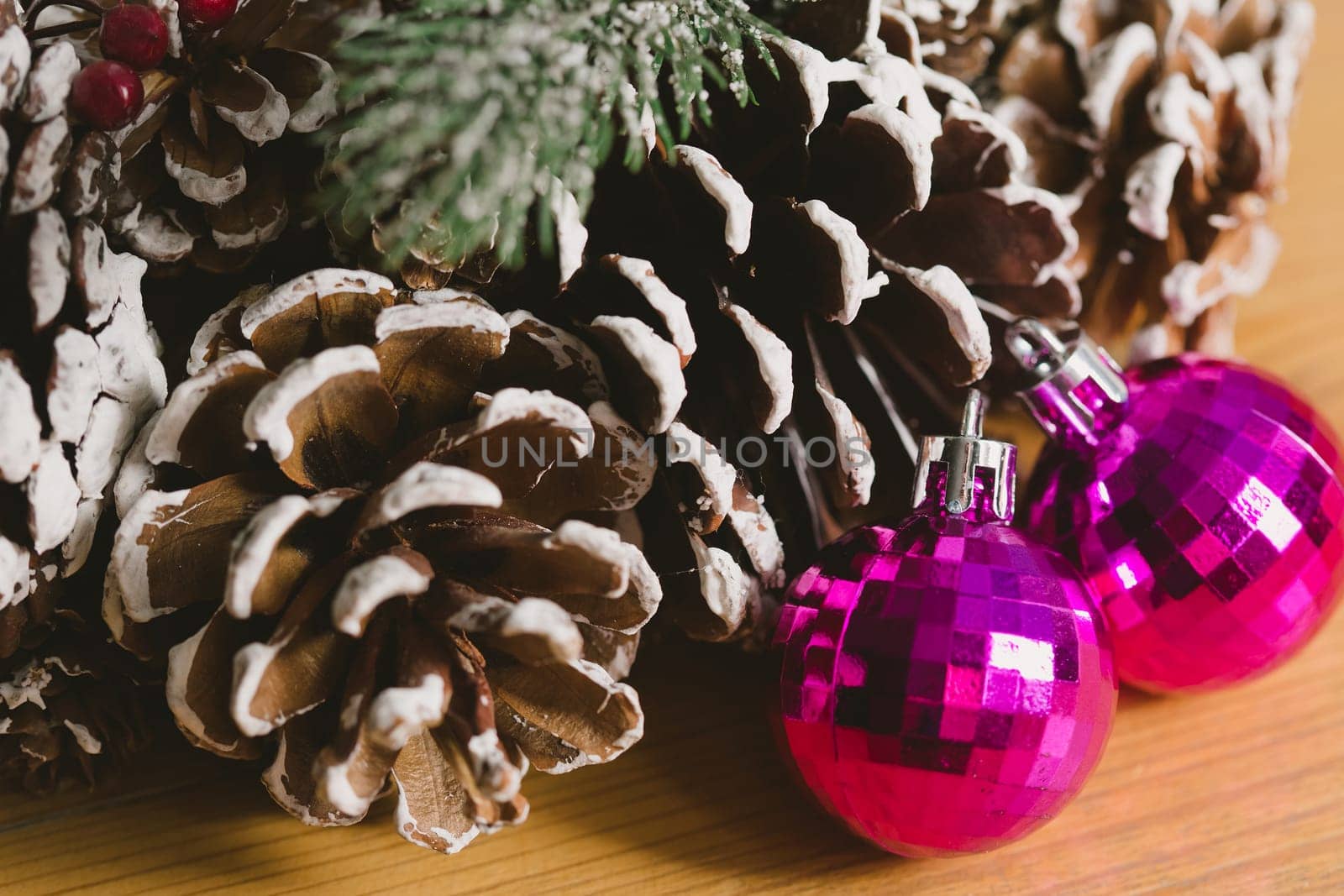 Holiday wreath adorned with snow-dusted pine cones, evergreen branches, red berries, and two shiny pink Christmas ornaments