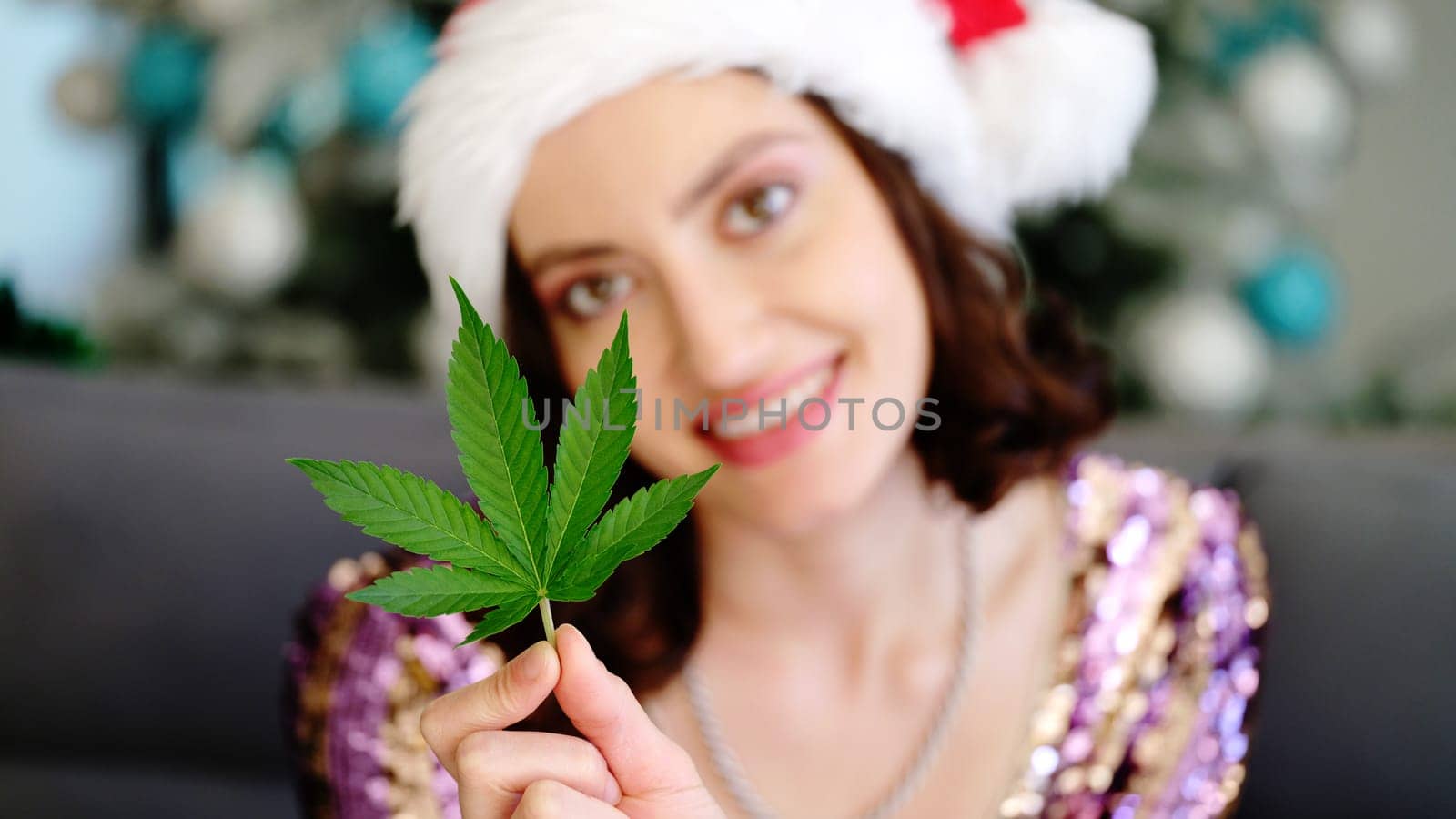 A cheerful woman in a Santa hat holds a vibrant green cannabis leaf among colorful holiday decorations