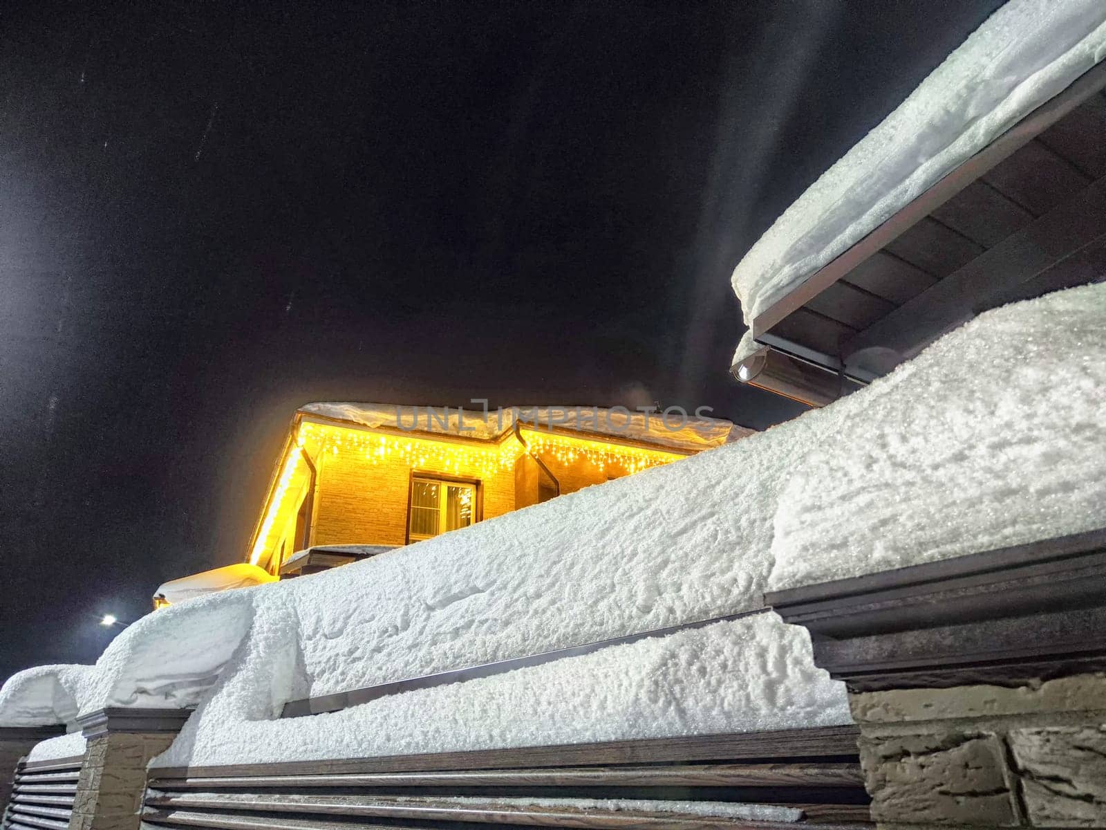 House decorated with bright Christmas garlands, surrounded by snow, under dark night sky