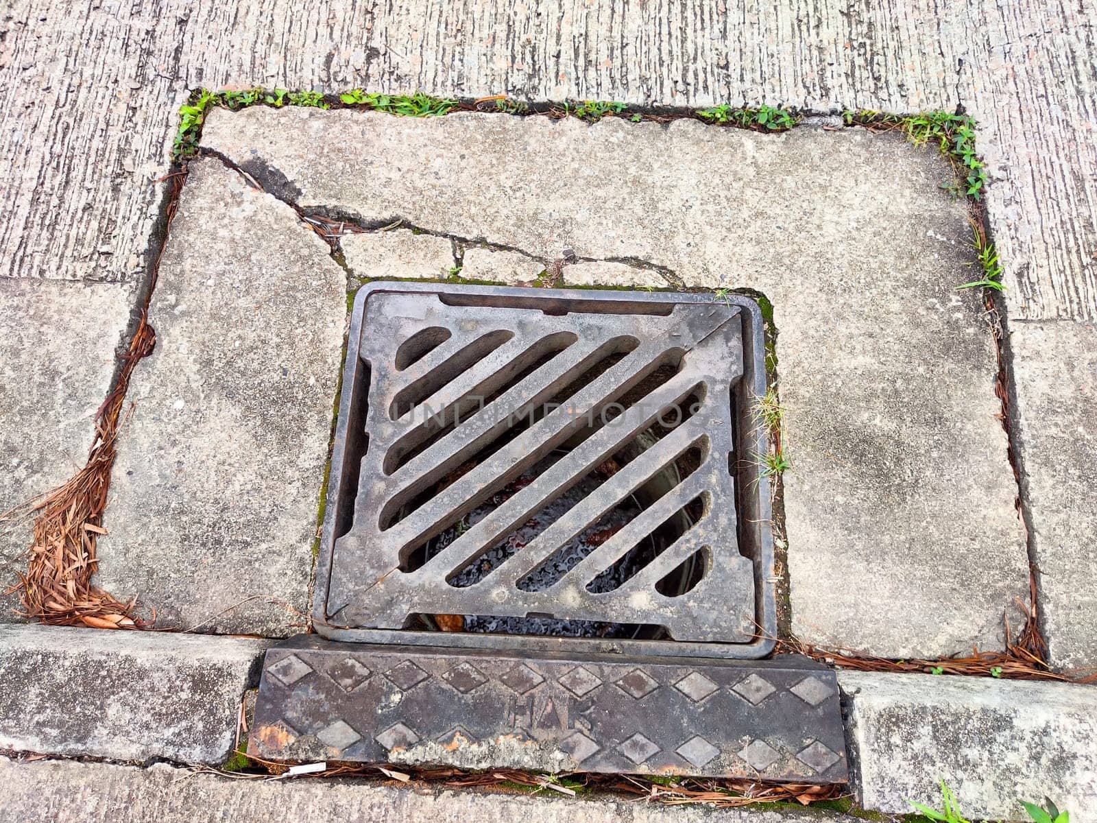 The cracked concrete sidewalk leads to a metal drainage cover filled with leaves, showing signs of neglect and environmental wear.