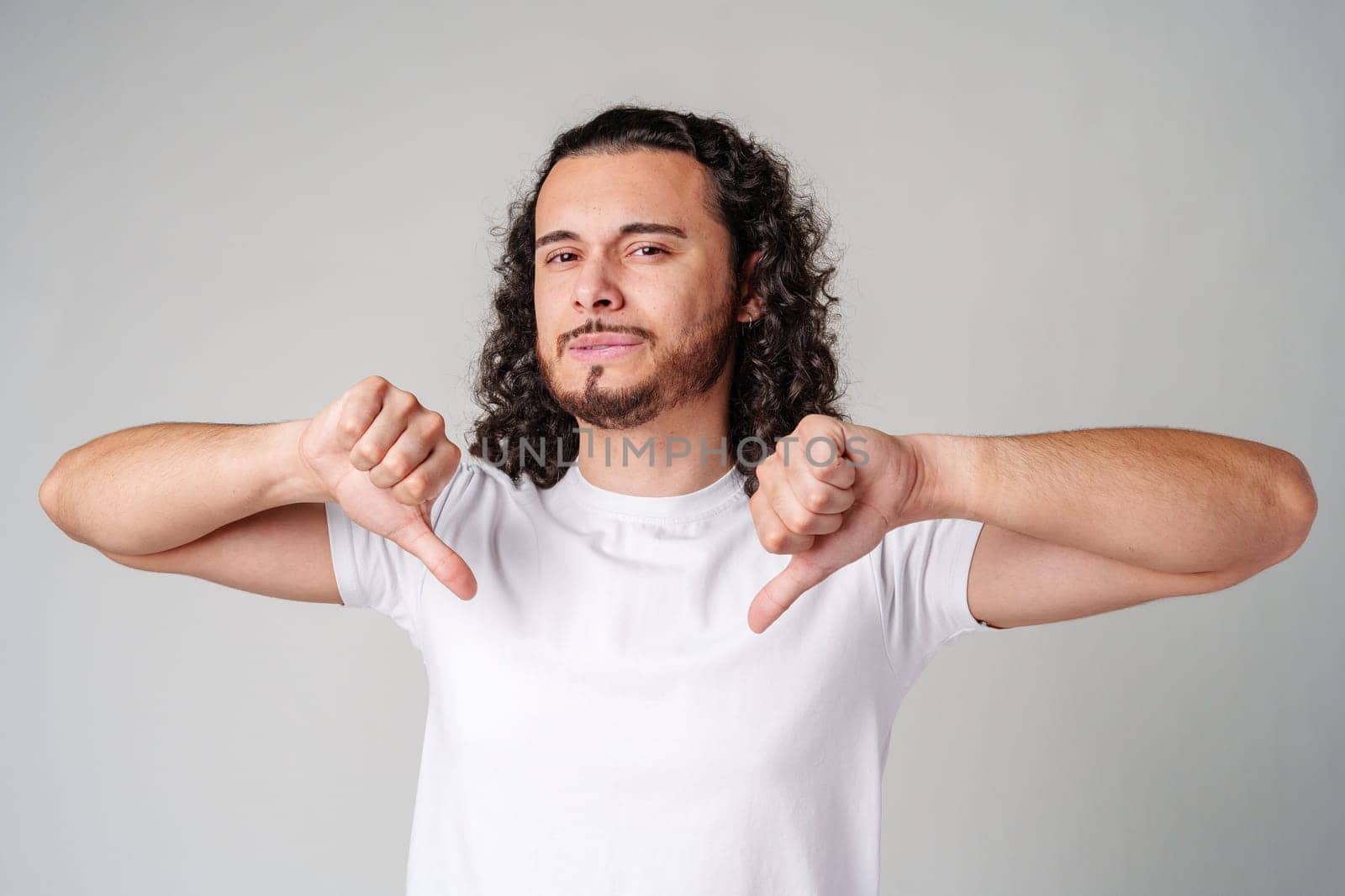 A young man with curly hair joyfully pointing down while wearing a plain white t-shirt against a neutral background close up