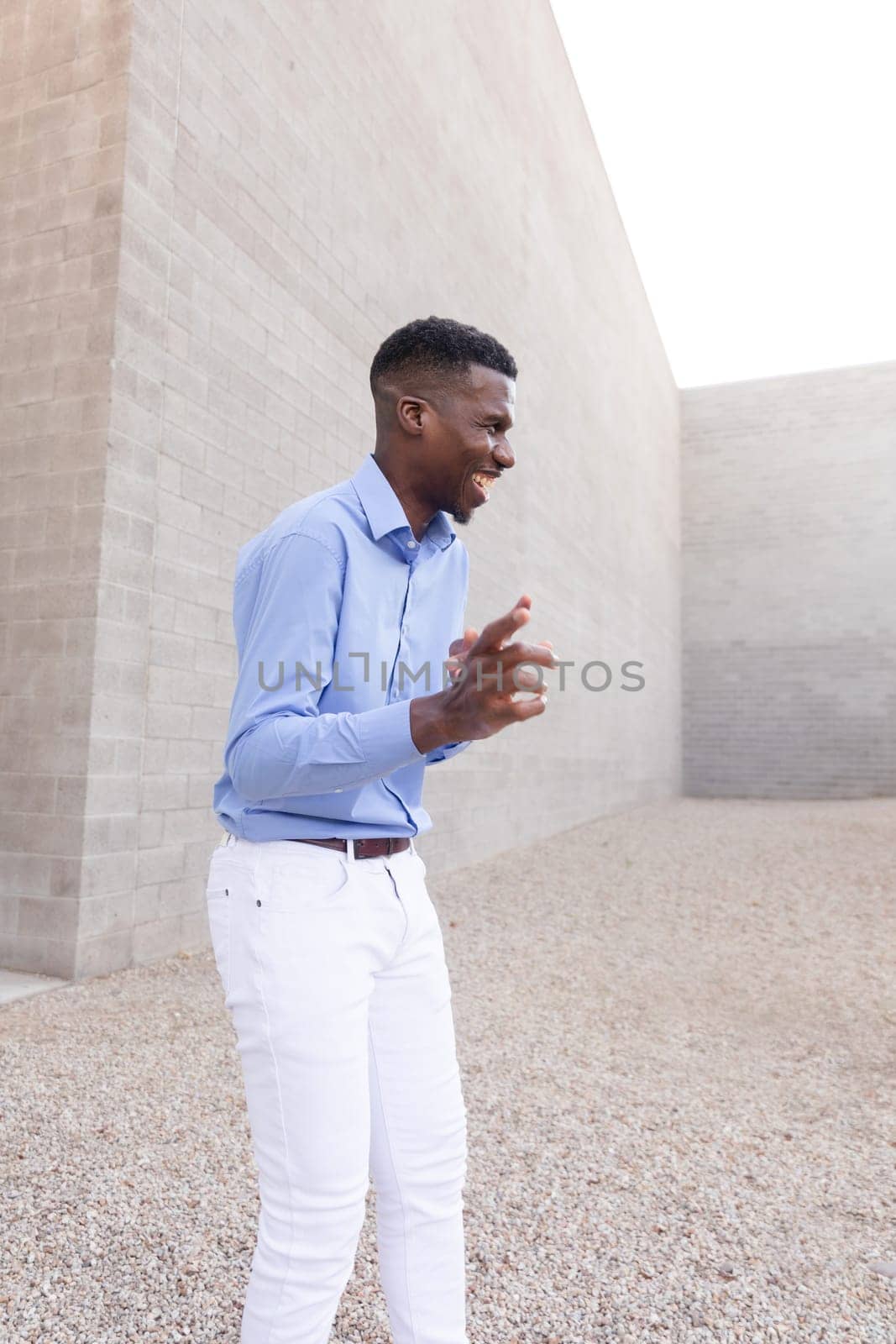 Laughing Stylish African American Man wearing White Jeans and Blue Shirt Outdoor, Concrete Wall on Background. Fashionable Middle Age Male in His 40s Portrait of Positive Ivorian. Vertical Plane