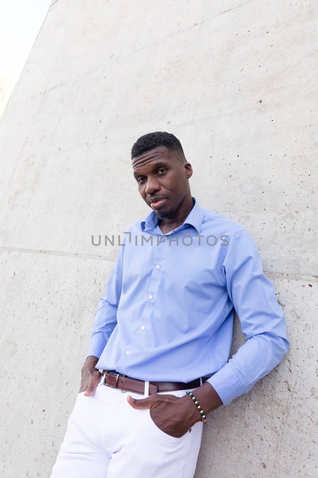 Portrait Of Handsome Stylish African American Man wearing White Jeans and Blue Shirt, Brown Belt Outdoor, Concrete Wall on Background. Fashionable Middle Age Male in His 40s,Ivorian. Vertical Plane