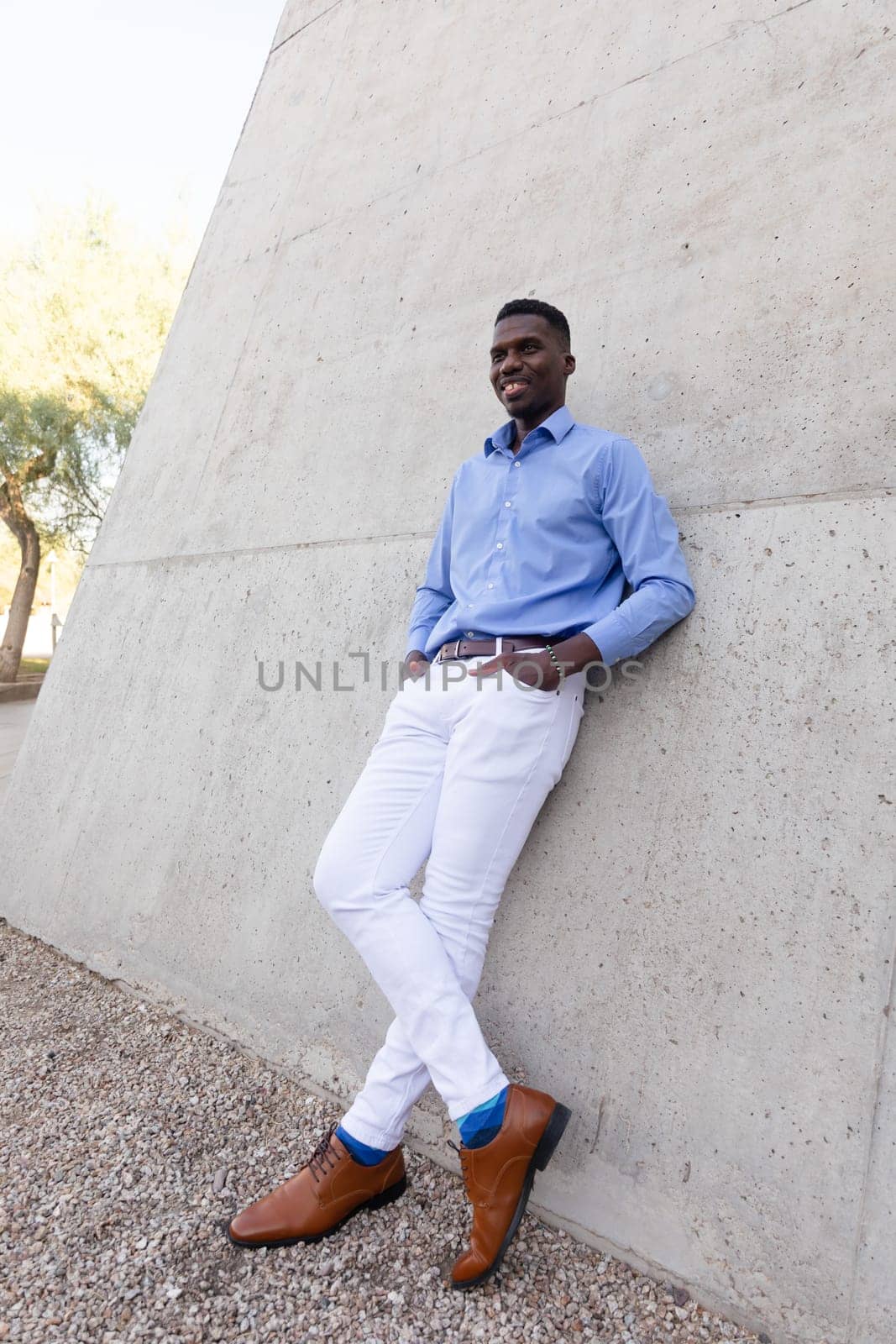 Full Size Handsome Stylish African American Man wearing White Jeans and Blue Shirt, Brown Belt, Shoes Outdoor, Concrete Wall on Background. Fashionable Middle Age Male in His 40s, Ivorian. Vertical.