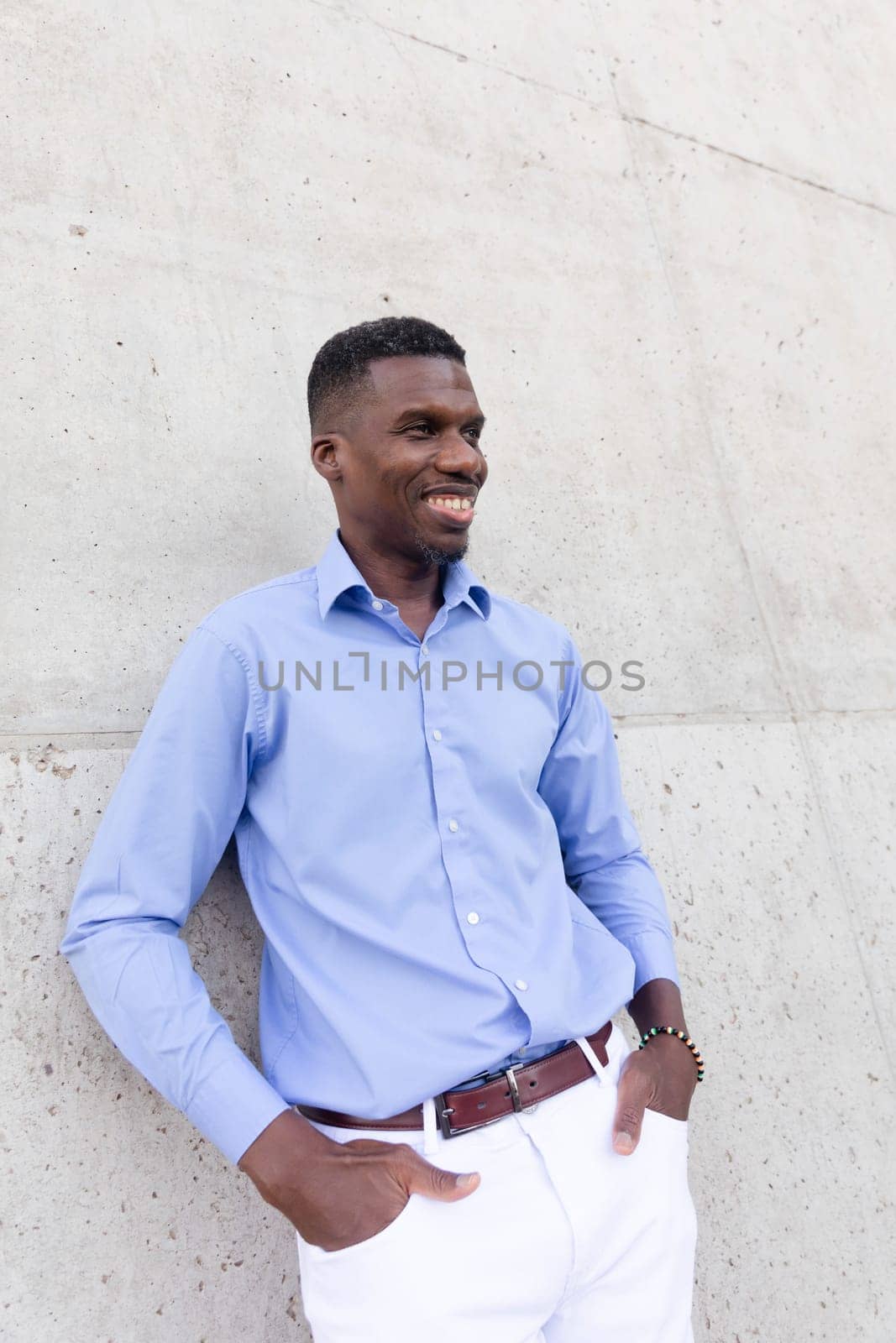 Portrait Of Handsome Smiling With Teeth African American Man wearing White Jeans and Blue Shirt, Brown Belt Outdoor, Concrete Wall on Background. Fashionable Male, Ivorian Vertical Plane