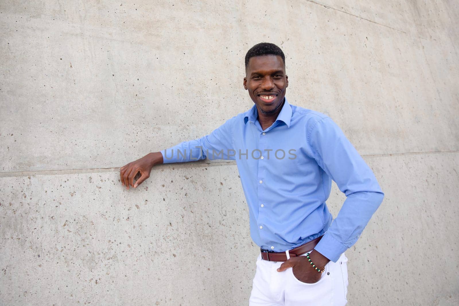 Portrait Of Handsome Smiling With Teeth African American Man wearing White Jeans and Blue Shirt, Brown Belt Outdoor, Concrete Wall on Background. Copy Space Fashionable Male, Ivorian. Horizontal.