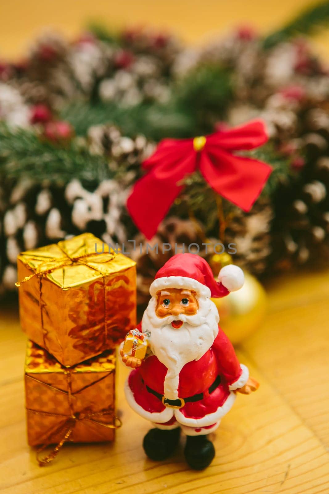 Christmas wreath decorated with pine cones, red bows, and evergreen branches sits on a wooden surface. In front of the wreath, a small Santa Claus figurine and golden ornaments add a cheerful holiday touch