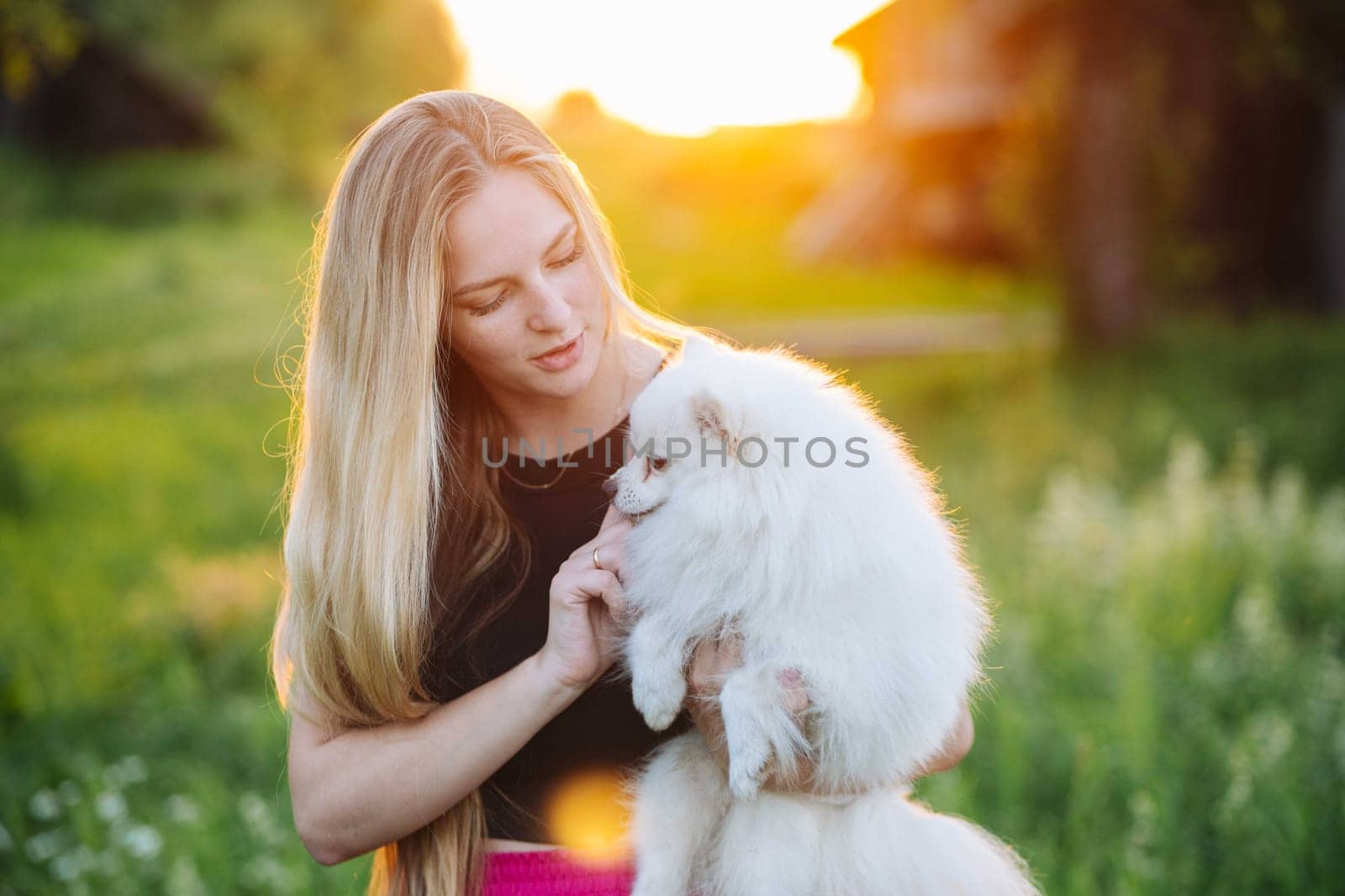 Blonde woman tenderly embraces her fluffy white dog in a vibrant field at sunset, exuding warmth and joy in the great outdoors