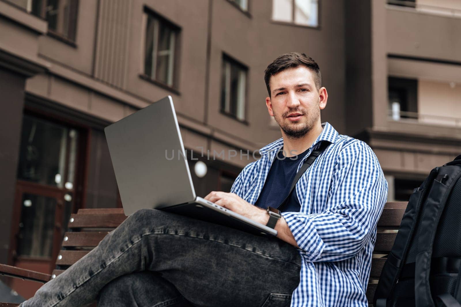 Young man working on a laptop while seated on a bench outside close up