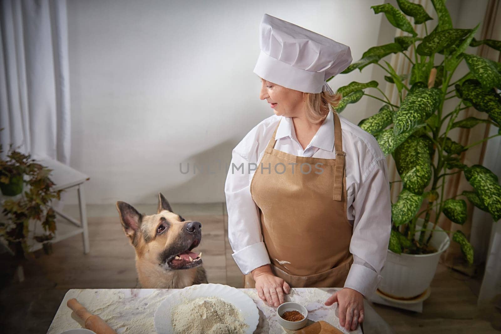 A cheerful woman in a chef uniform mixes flour while her eager shepherd dog waits for a tasty morsel, capturing a playful cooking moment.
