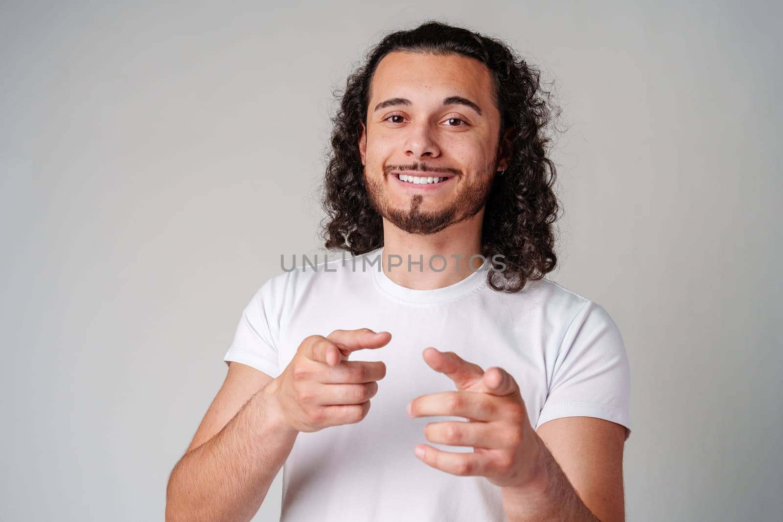A smiling young man with curly hair playfully pointing directly at the viewer in a light-colored setting close up