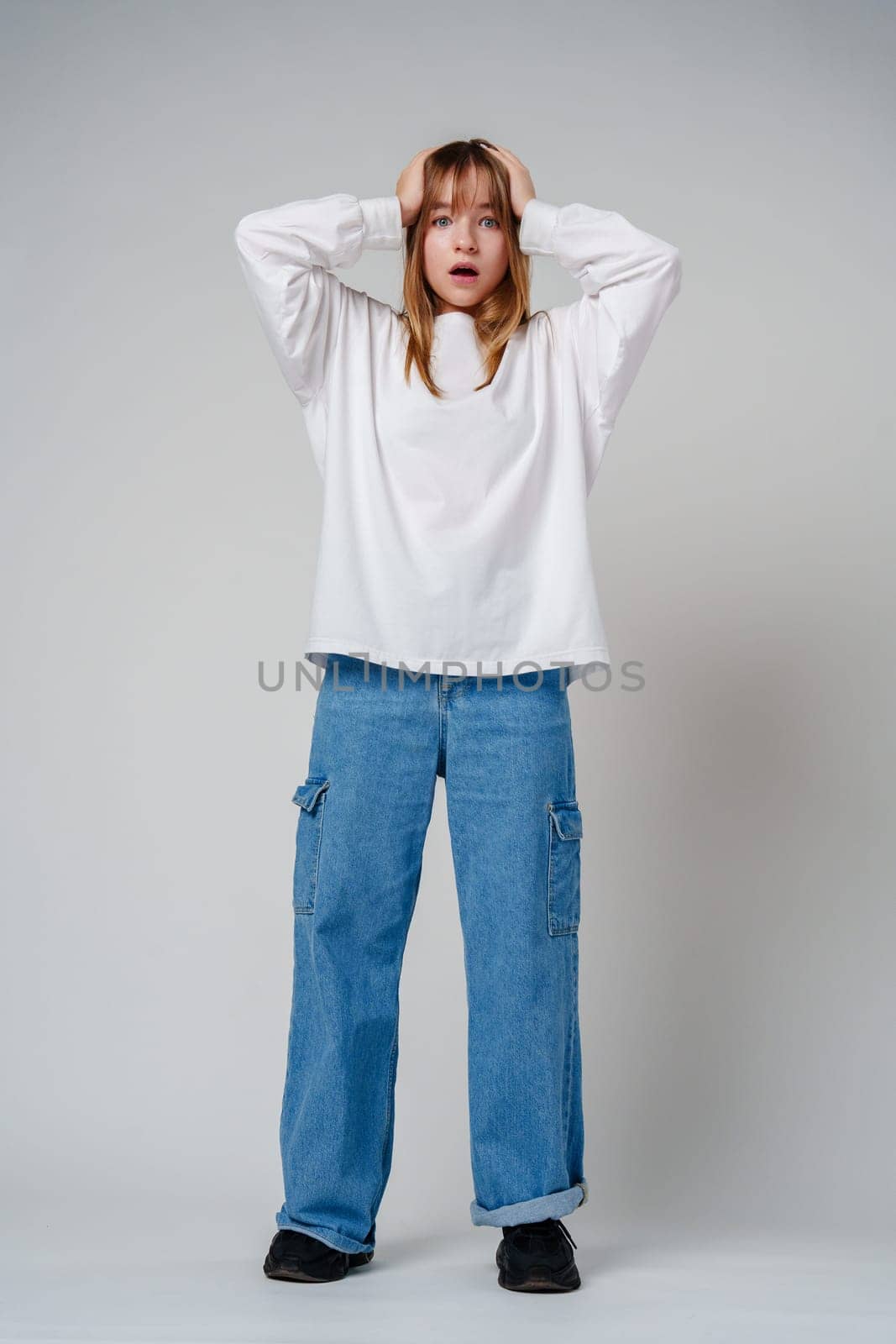 A teen girl in a loose white sweater looks surprised with her hands on her head against a plain gray backdrop close up