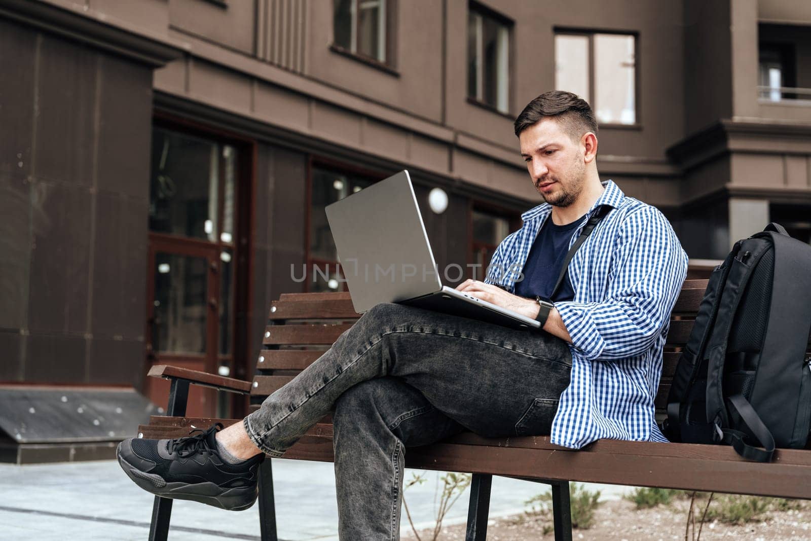 Young man working on a laptop while seated on a bench outside close up