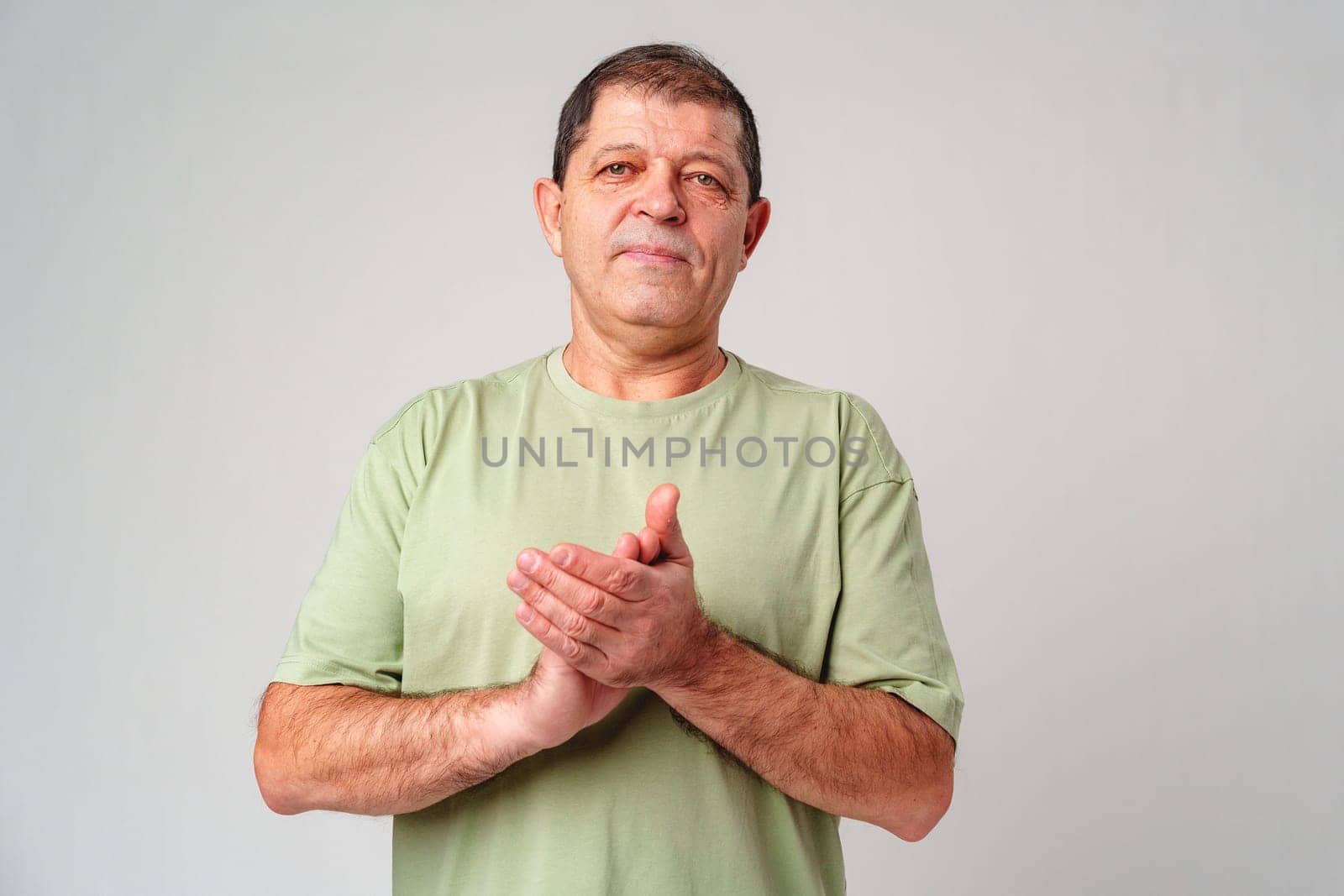 Elderly man claps his hands while standing against a neutral background in a casual light green shirt close up