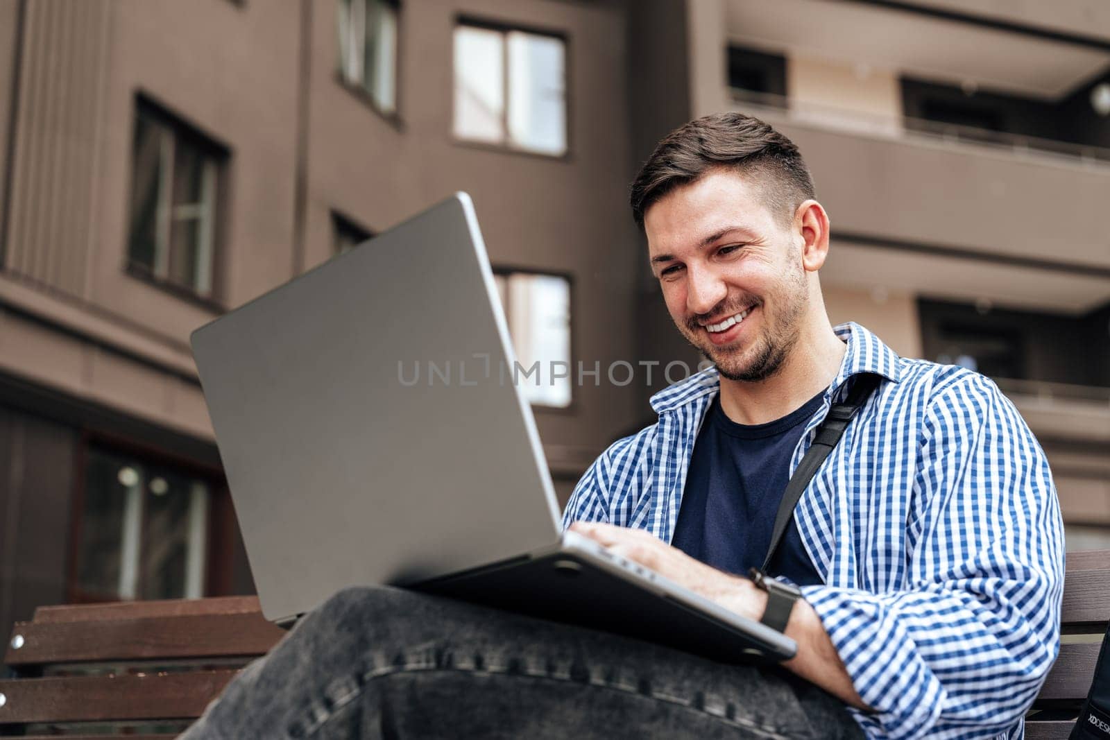 Young man working on a laptop while seated on a bench outside close up