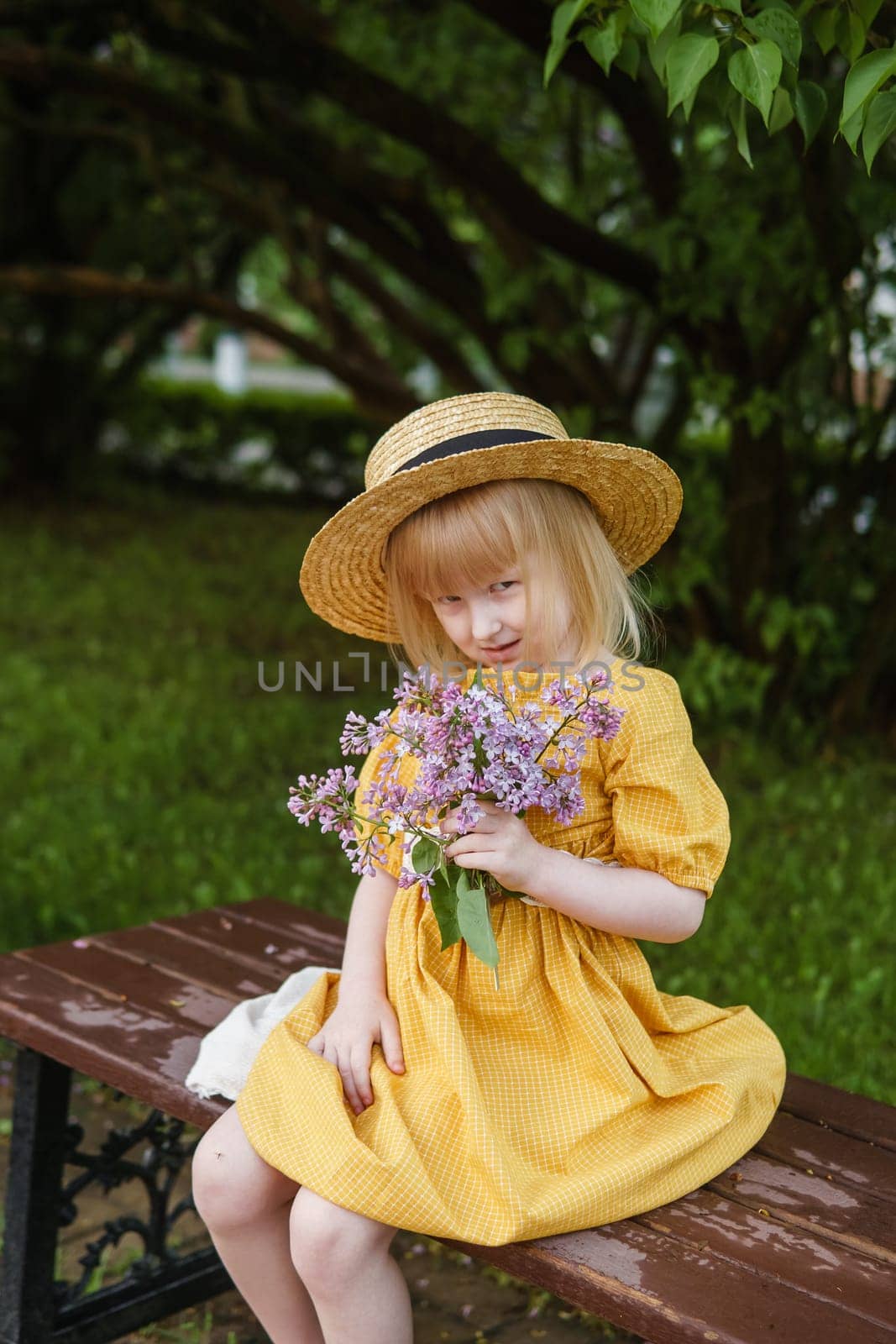 A little girl in a yellow dress and straw hat wearing a bouquet of lilacs. A walk in a spring park, blossoming lilacs