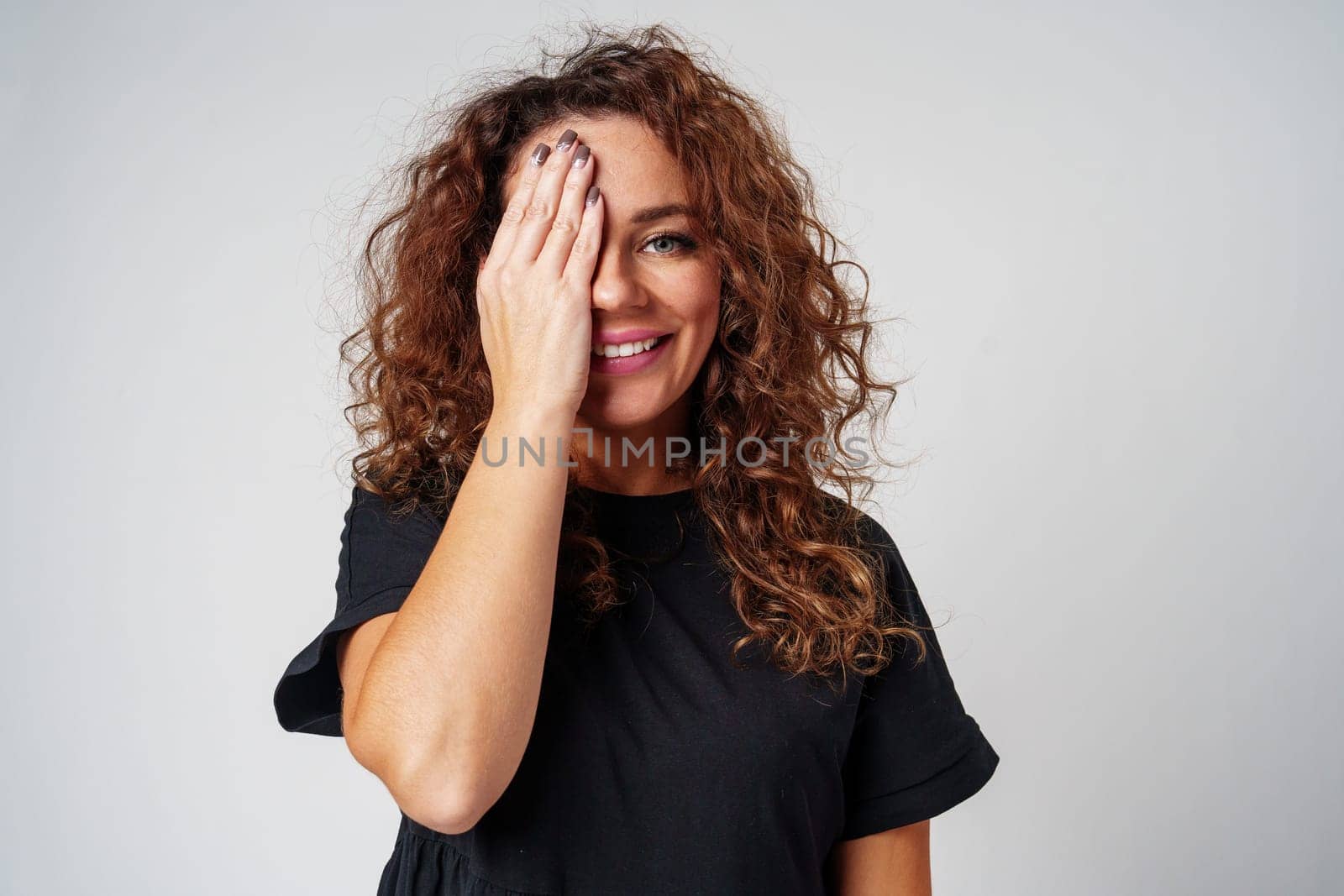 A woman with curly hair playfully covering one eye, smiling, against a neutral background