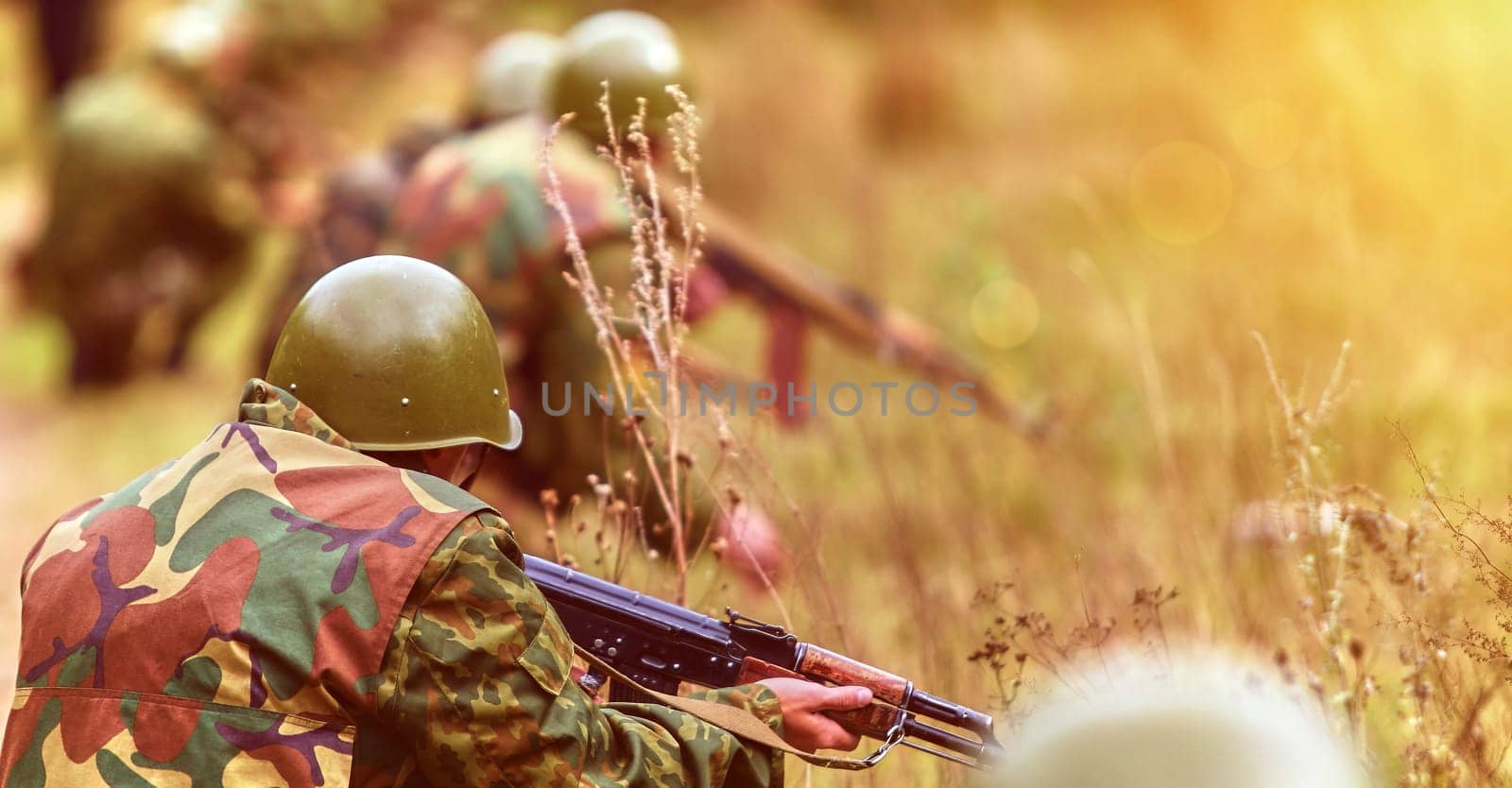 A group of soldiers crouches in a grassy area, gripping their rifles as they prepare for drills. The early morning light highlights their focused expressions and camouflage attire.
