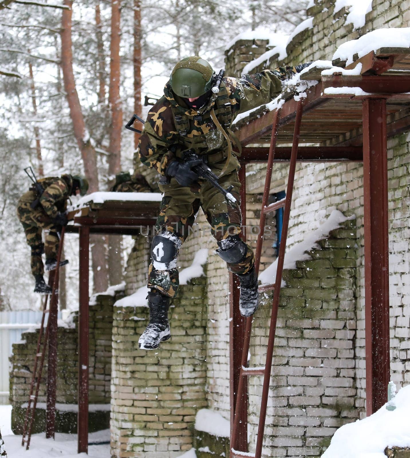 Special forces soldiers climbing a building in combat gear during a military exercise. Armed with rifles, helmets, and protective gear. Brick building with windows in the woods.