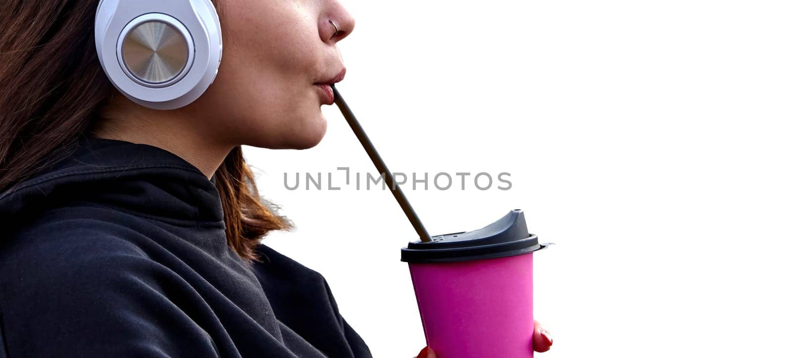 A young woman with headphones takes a sip from a pink coffee cup. She appears relaxed and content, enjoying her beverage in the fresh air.
