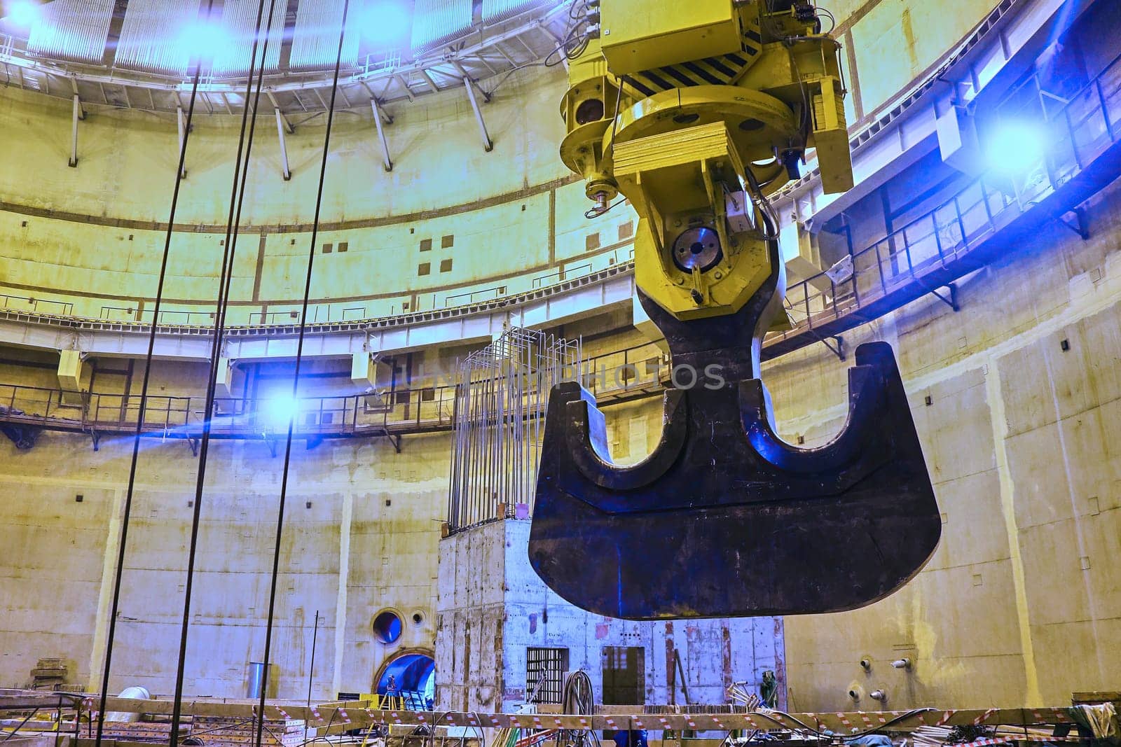 A large, steel claw hangs suspended from a crane inside a vast, concrete structure. The claw appears to be reaching towards a shaft of sunlight that illuminates the room. The structure appears to be under construction and is surrounded by scaffolding and other building materials.