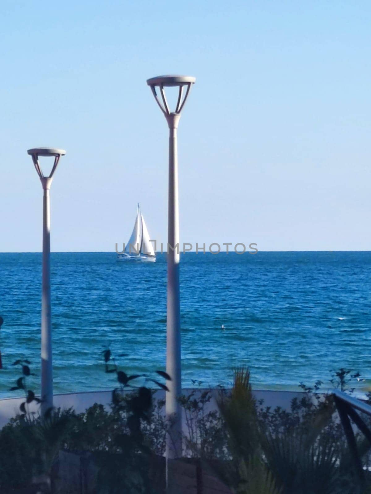 Vertical seascape, street lights on the embankment against the backdrop of the sea horizon and sailboat.