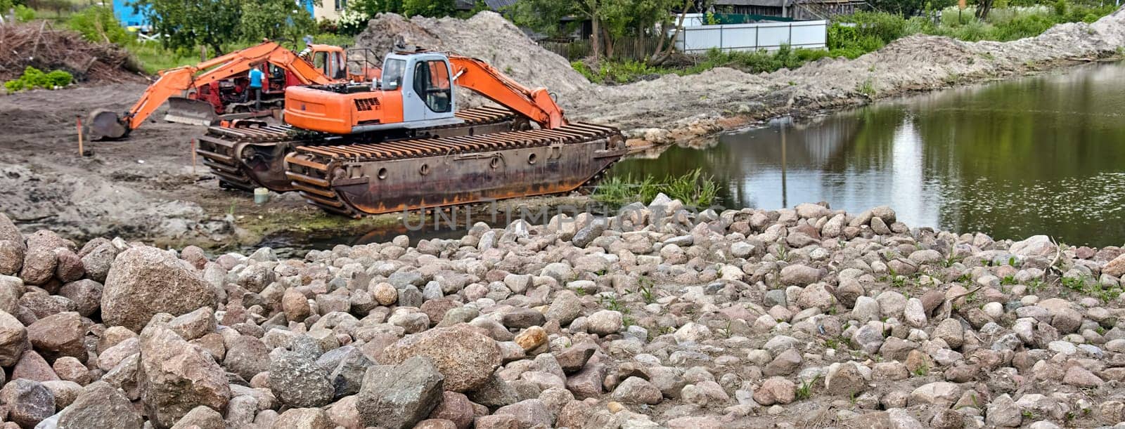 An orange and gray amphibious excavator works on the edge of a pond, placing large rocks to stabilize the shoreline. The excavator's tracks are partially submerged in the water.