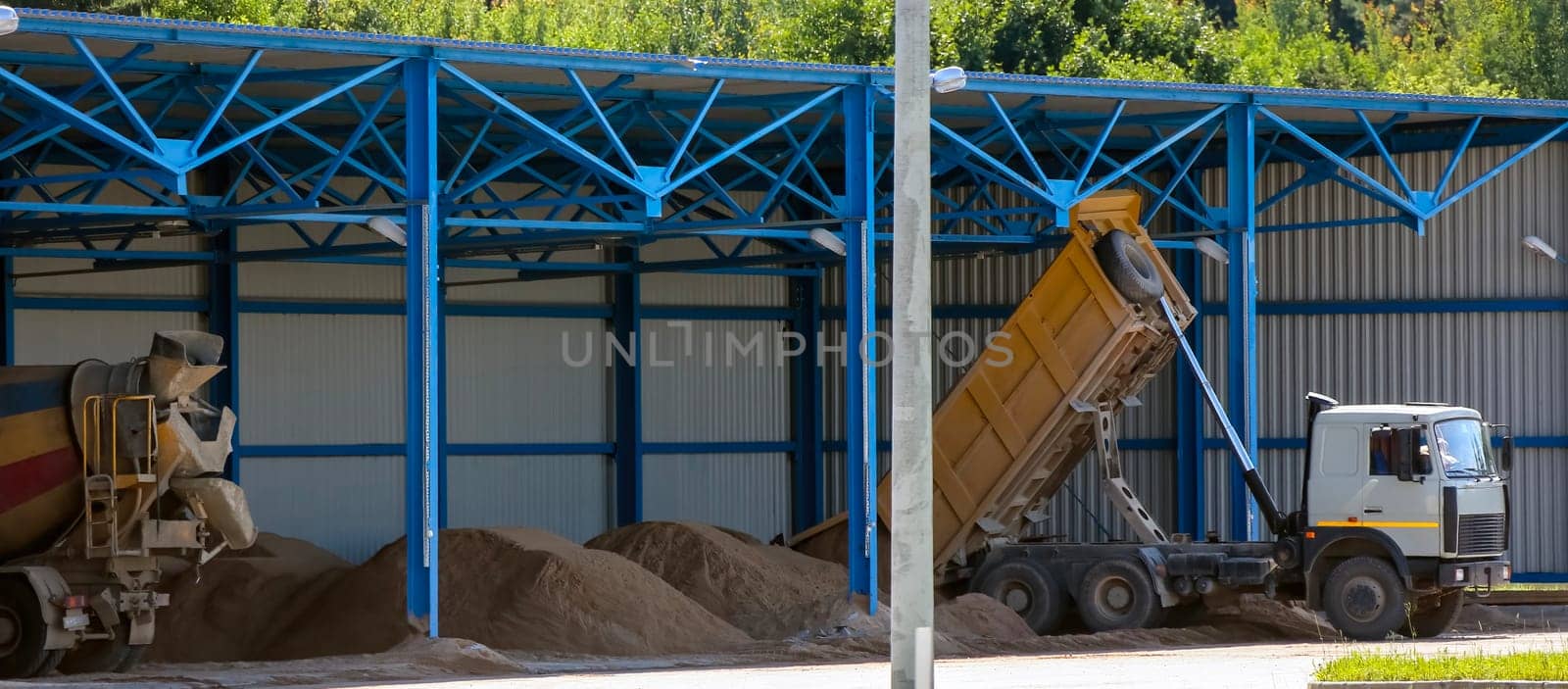 In a bustling construction area, trucks unload their cargoes of sand and gravel under bright blue skies. A large yellow dump truck raises its bed, releasing a cascade of material into a designated space. Nearby, concrete mixers wait patiently, their large compartments filled with a mixture prepared for upcoming projects.