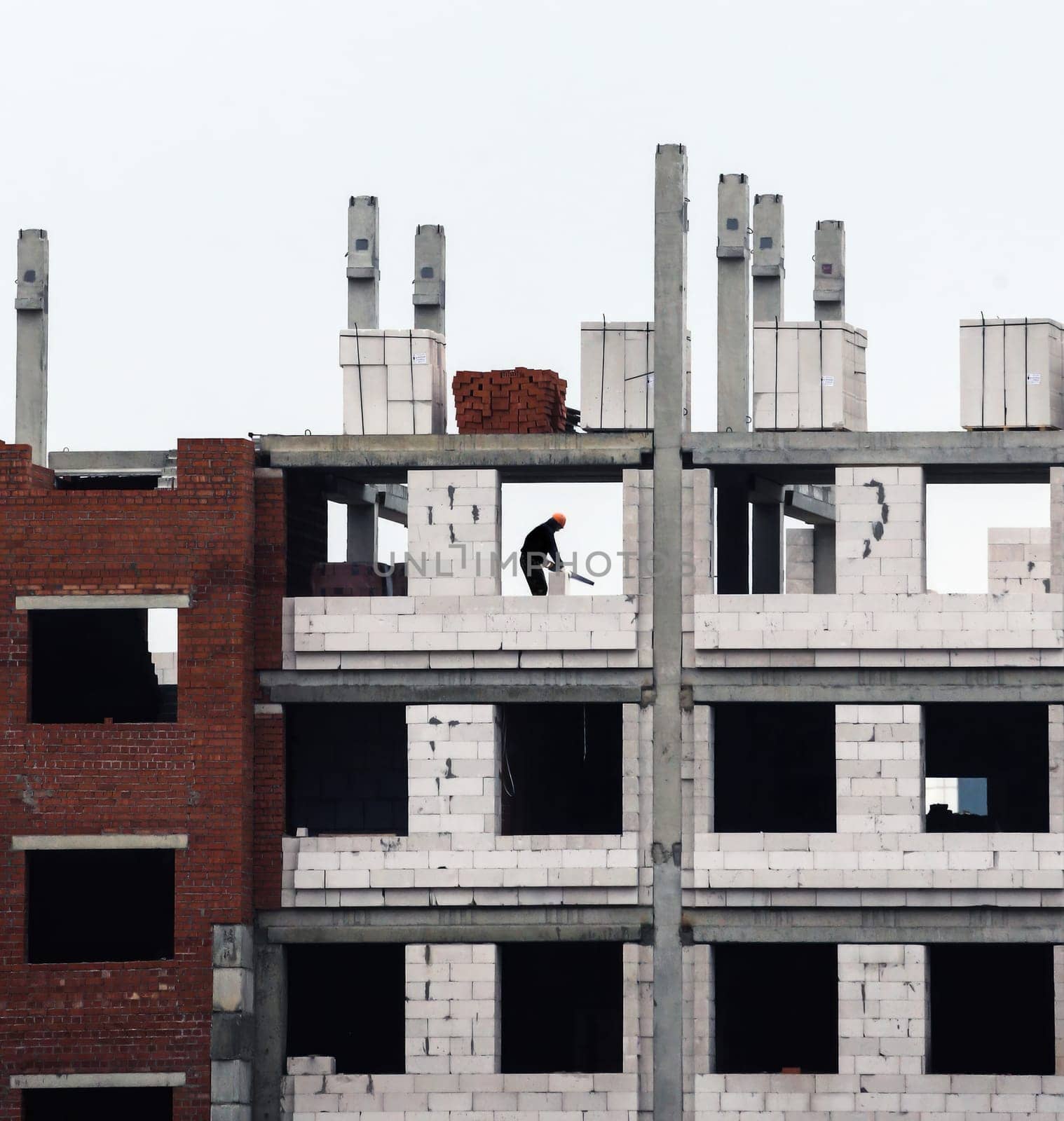 A solitary worker is seen meticulously laying bricks on the scaffolding of a rising multi-story building. Surrounding him are the skeletal remains of the structure, showcasing unfinished concrete and brickwork. Above, gray clouds hang low, hinting at impending rain, while the morning light filters in, casting shadows across the building.