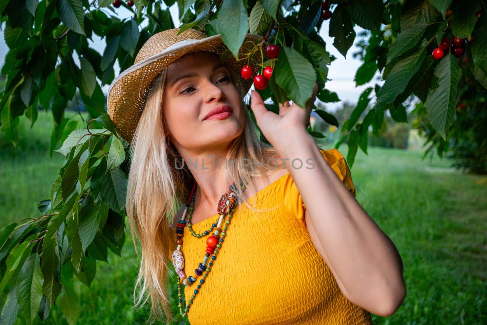 A cheerful blonde woman in a mustard dress stands under a cherry tree holding ripe red cherries. The lush green leaves and warm spring sunlight complete the scene.