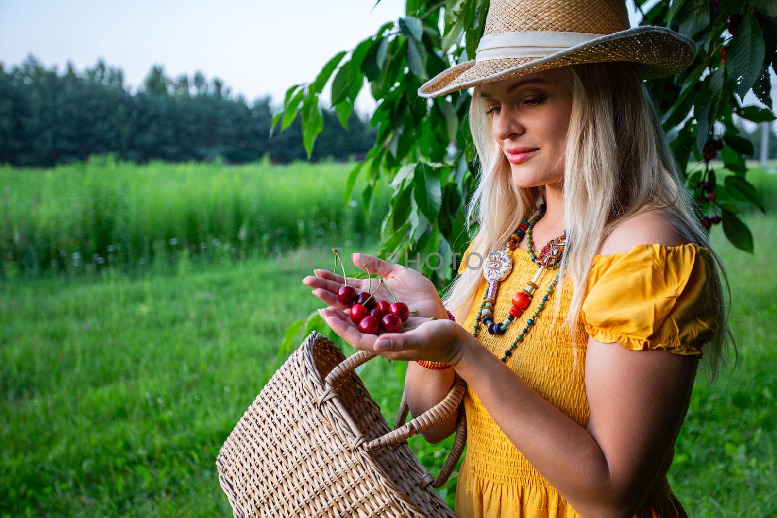 A smiling farmer in a mustard dress and hat holds ripe red cherries in her open hands. The close-up captures the cherries and a woven basket against a blurred grassy background.