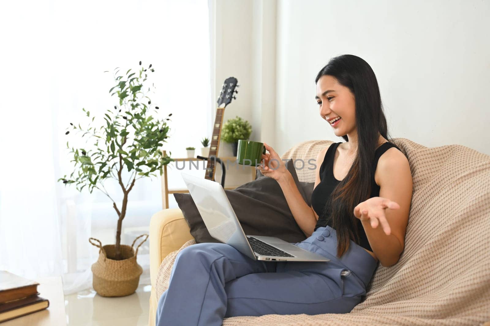 Smiling young woman having a casual video chat on laptop and drinking coffee on sofa.