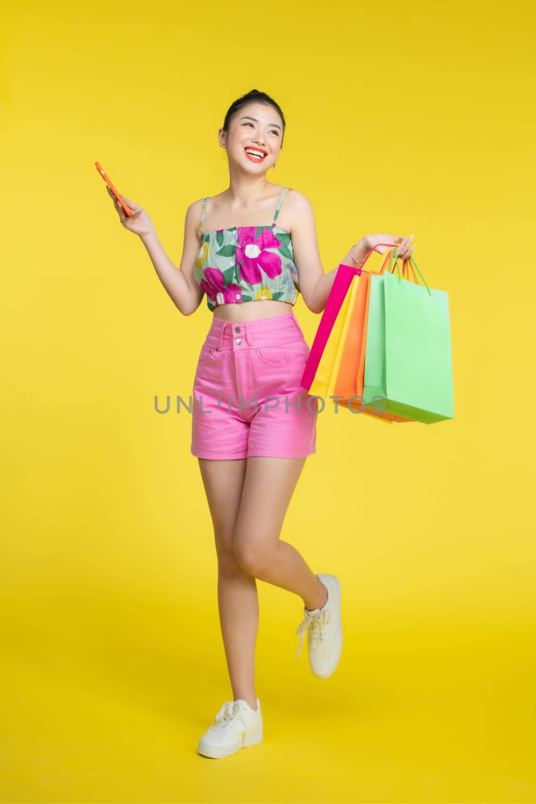Confident fashionable young Asian woman in sexy dress holding colorful shopping bags and smartphone