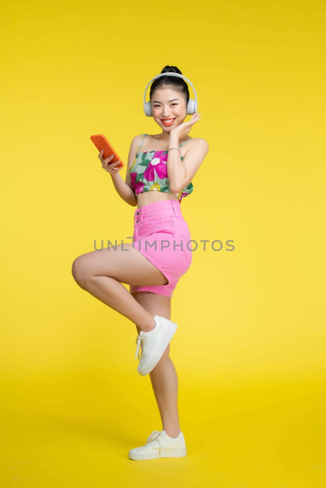 Happy smiling young Asian woman listening to music from headphones isolated on yellow background