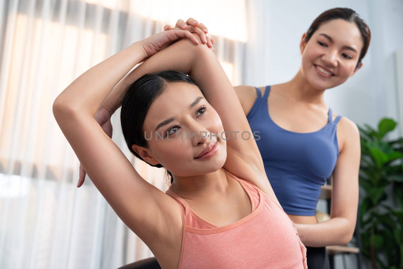 Asian woman in sportswear doing yoga exercise on fitness mat with trainer or workout buddy as home workout training routine. Healthy body care in yoga lifestyle people. Vigorous