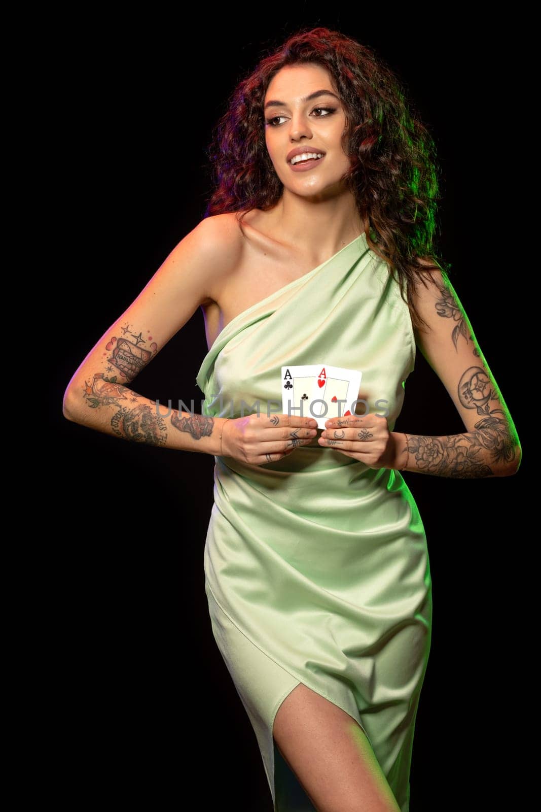 Smiling gambling attractive young woman in light green silk dress with tattoo on arm holding winning set of two aces cards posing against black background. Poker game concept