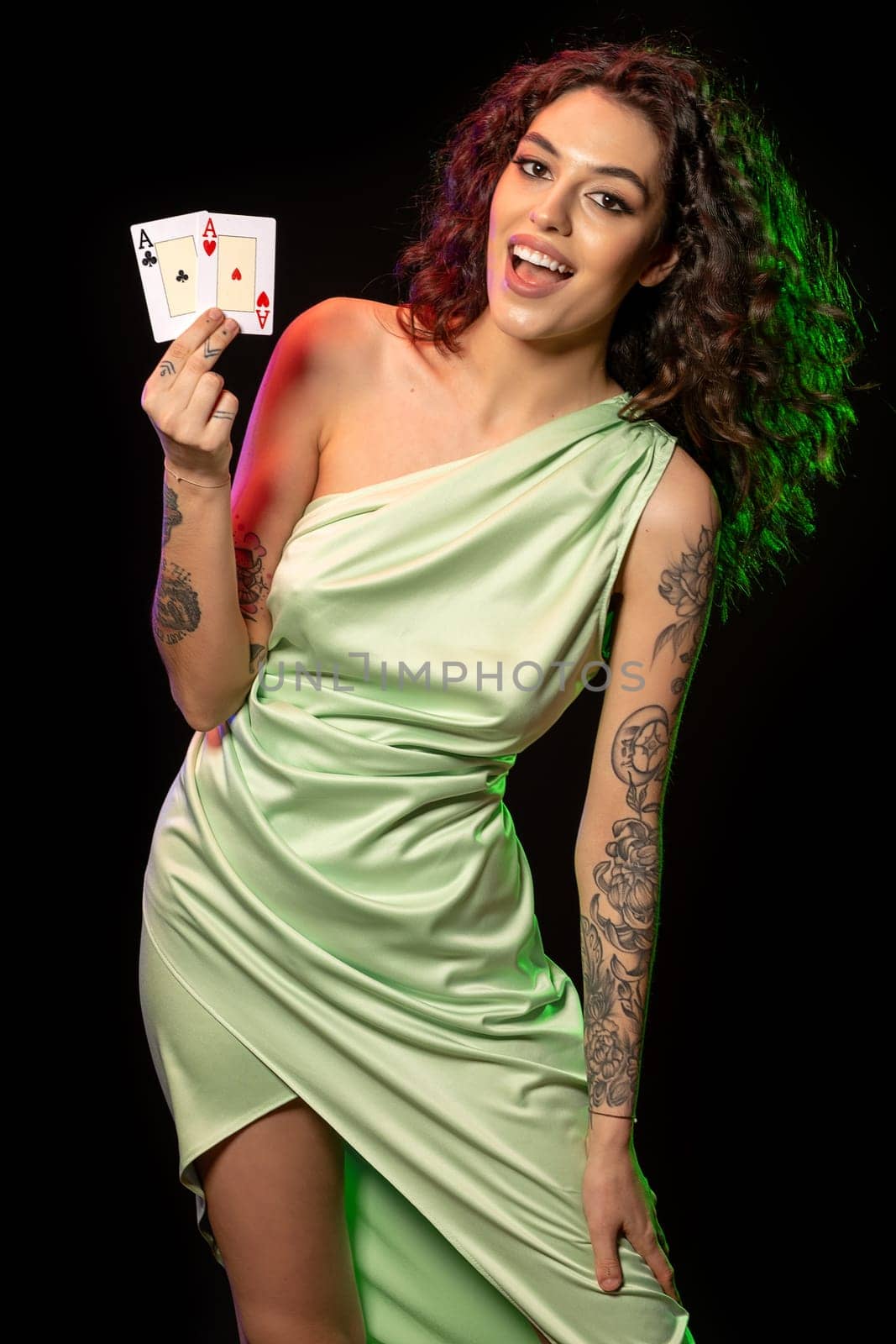 Cheerful stylish young woman with wavy dark hair wearing in silk light green dress standing against black background, holding set of winning cards of pair of aces. Successful gambling concept