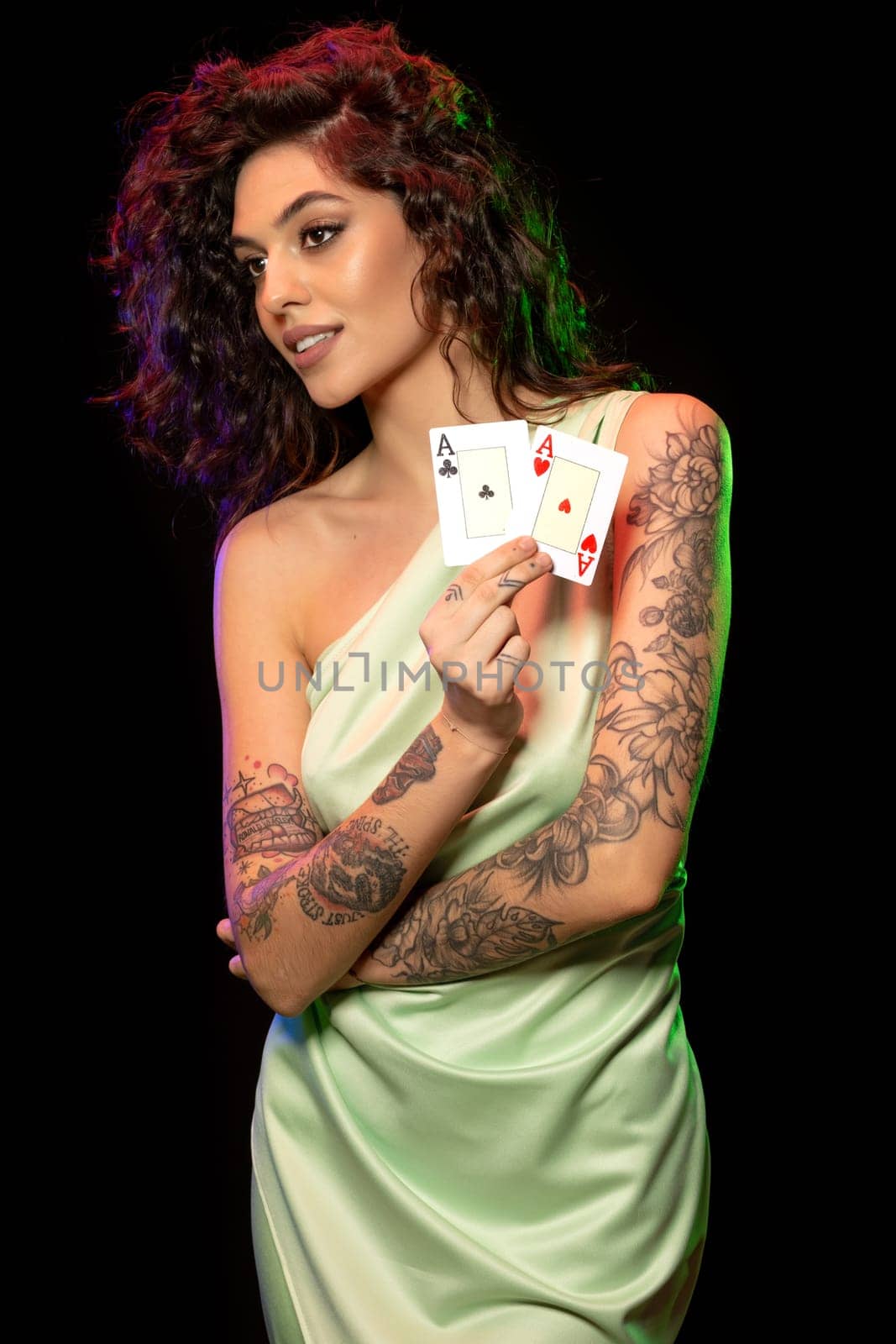 Lucky elegant young brown-haired girl in silk dress satisfied with successful poker game standing against black background, showing set of winning cards from pair of aces. Fortune smiled