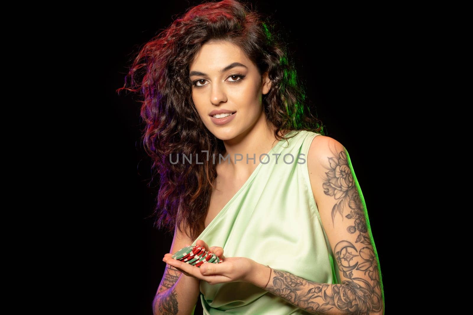 Attractive young dark-haired woman with tattoos on arms holding red and green betting tokens in palms, satisfied with winning poker game. Closeup portrait. Concept of successful gambling